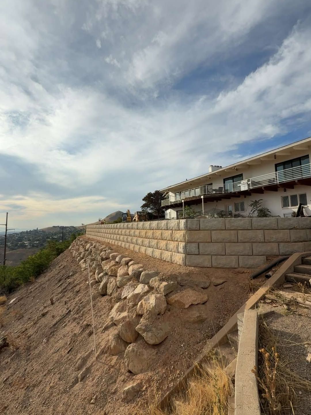 Retaining walls support a house built on a hillside; dry, rocky ground. Cloudy sky.