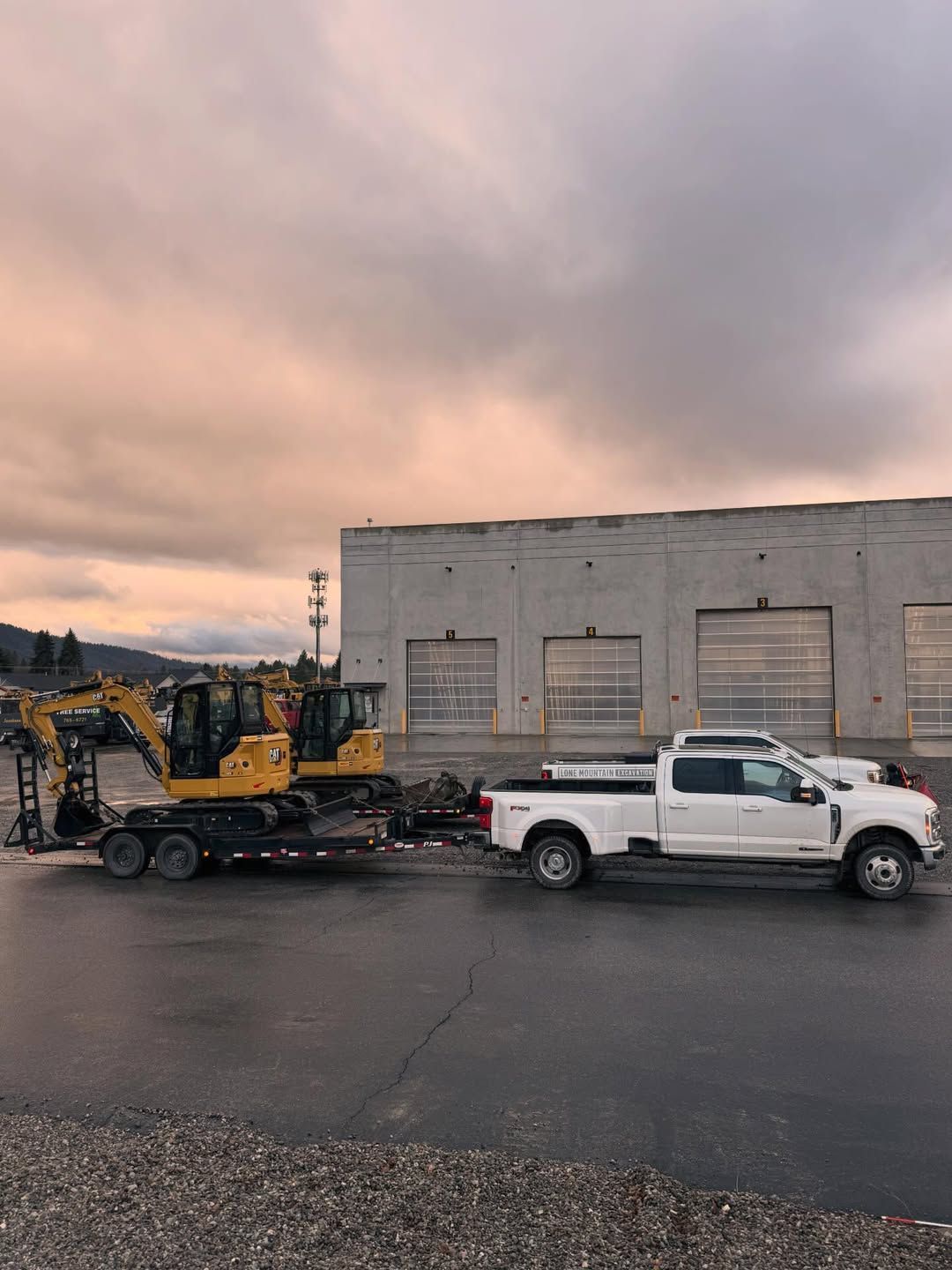 White pickup truck towing a yellow excavator on a trailer, in front of a gray building.