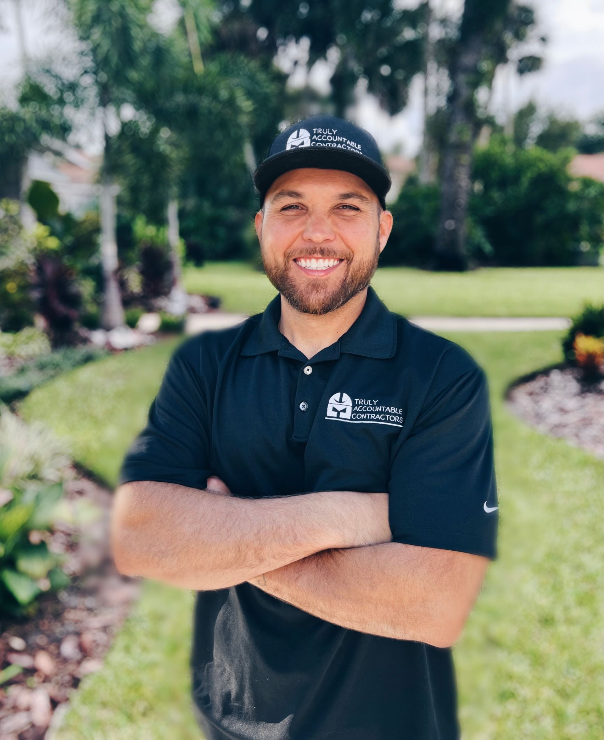 Man with crossed arms smiles, wearing a black polo shirt and hat, outdoors.