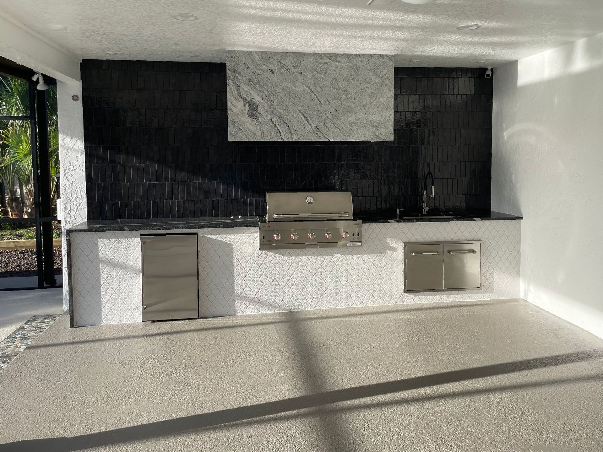 Outdoor kitchen with stainless steel grill, black tile backsplash, and white speckled countertop.