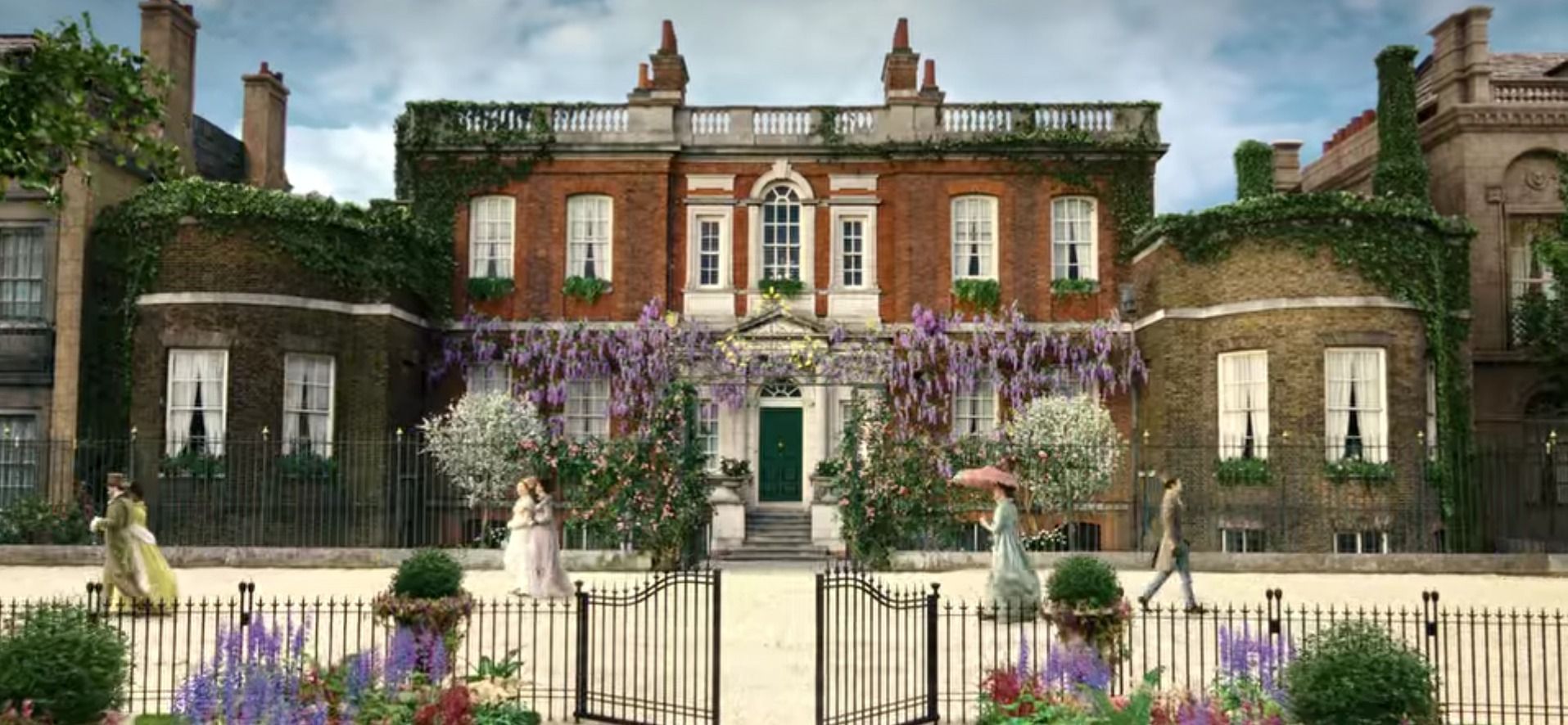 A large brick house with a green door is surrounded by flowers and a fence.