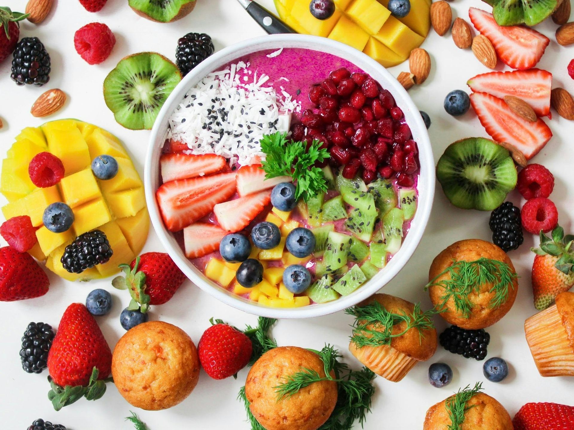 A bowl of fruit surrounded by muffins and berries on a table.