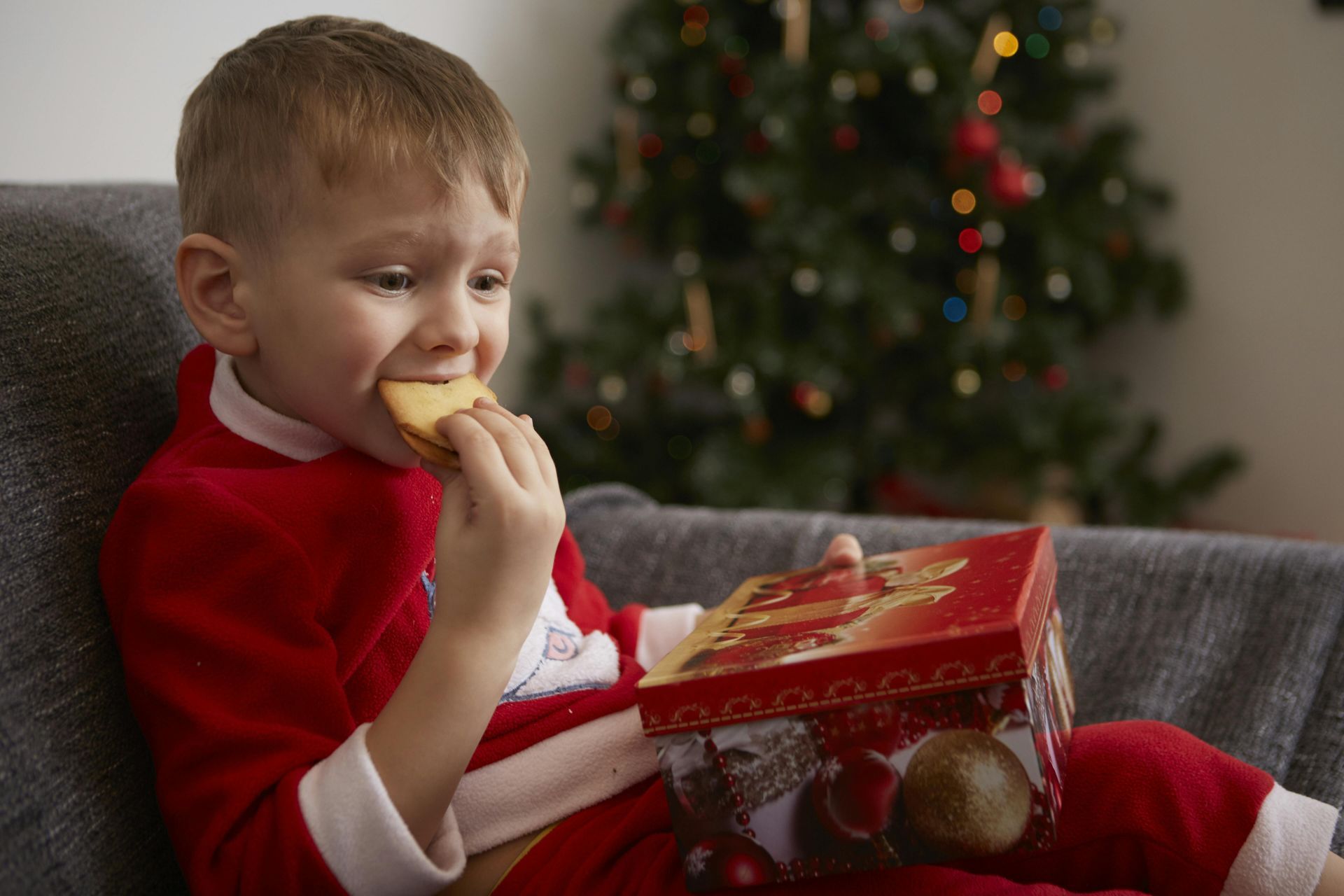 Boy in Santa pajamas eating a cookie from a decorated box on a sofa with a Christmas tree in the background.