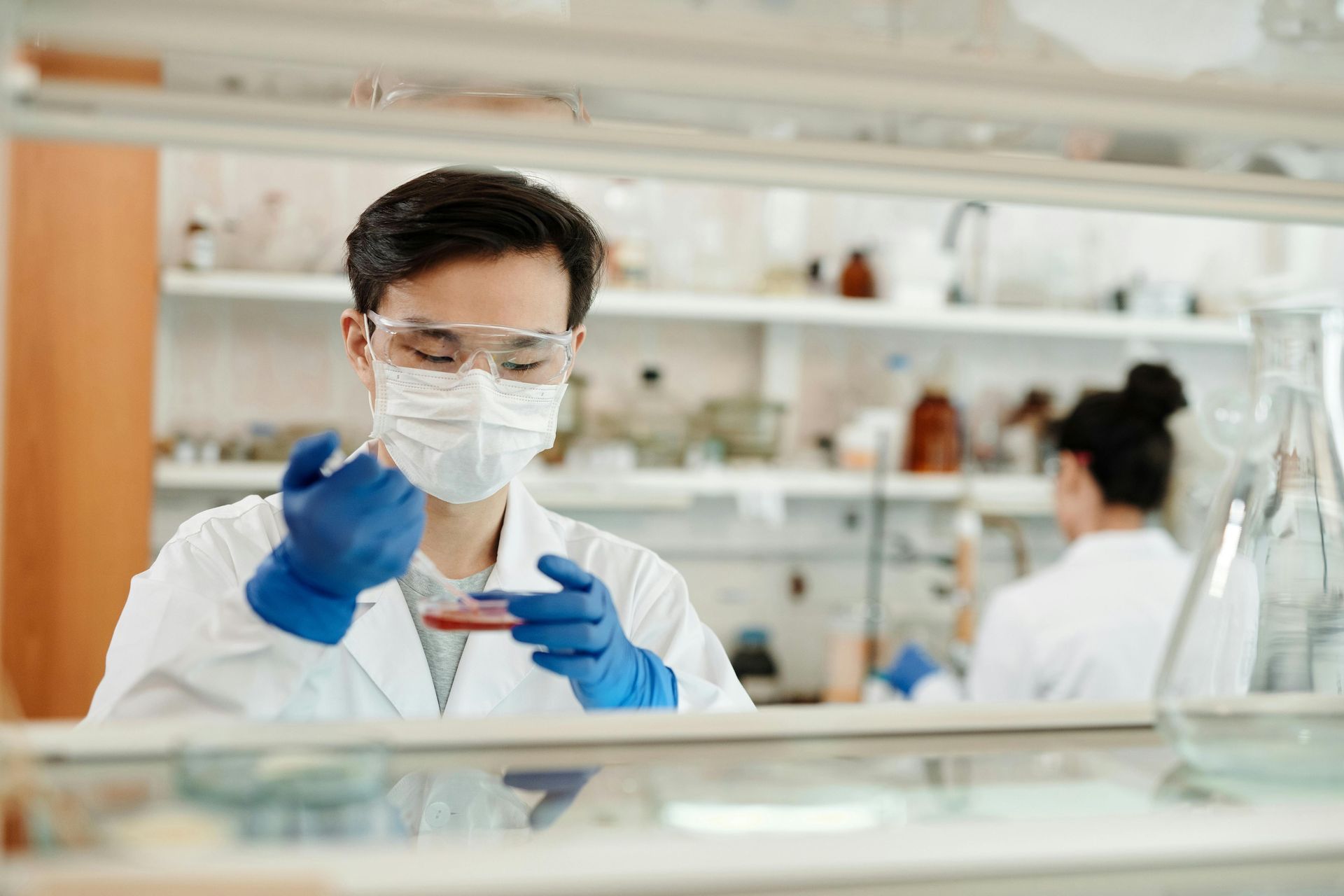 A scientist wearing a mask and goggles is holding a petri dish in a laboratory.
