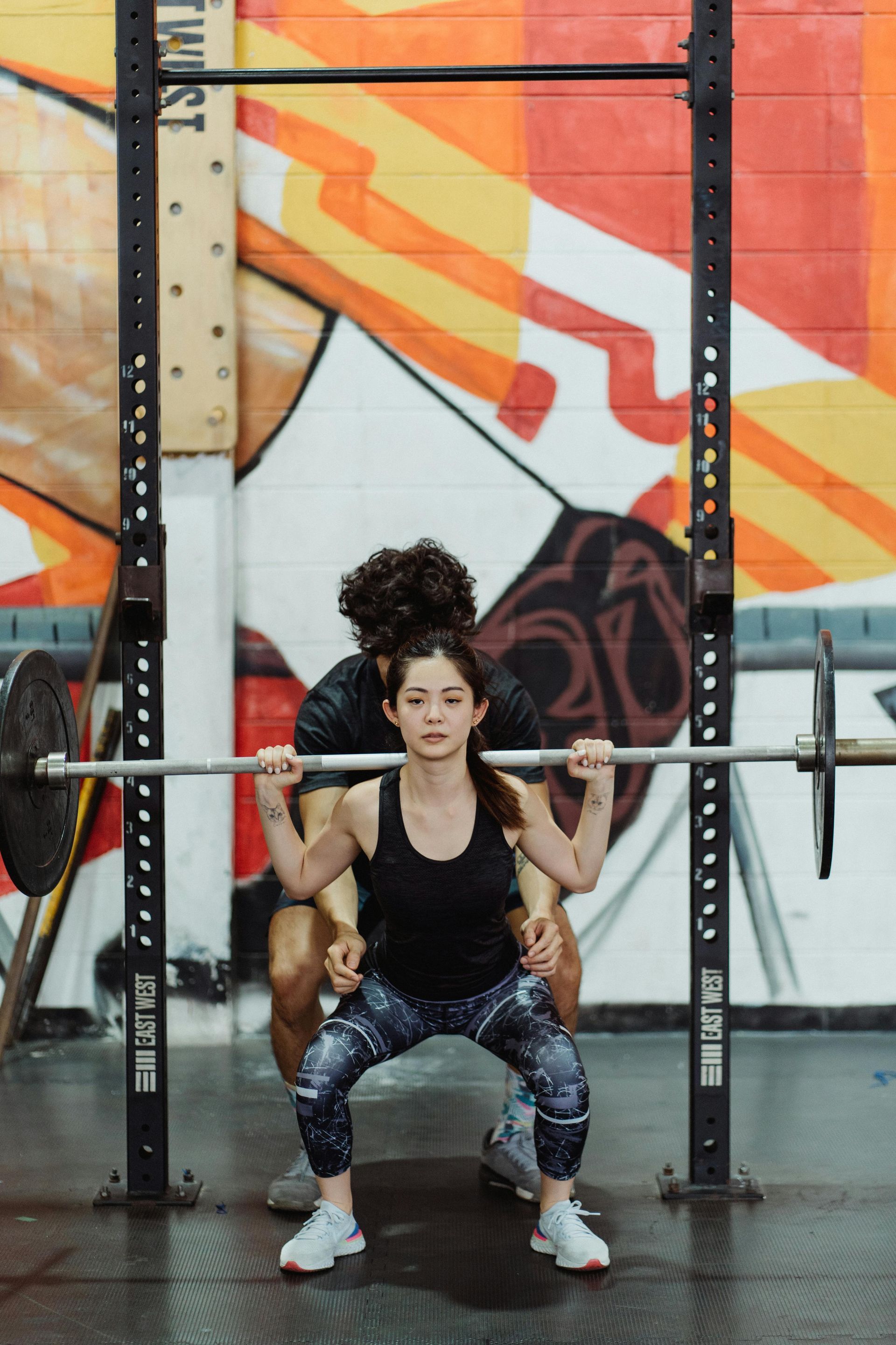 A man is helping a woman squat with a barbell in a gym.