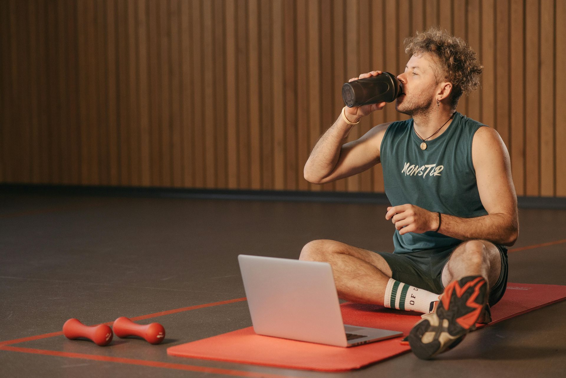 Man drinking from a shaker bottle while sitting on a mat with a laptop and dumbbells.