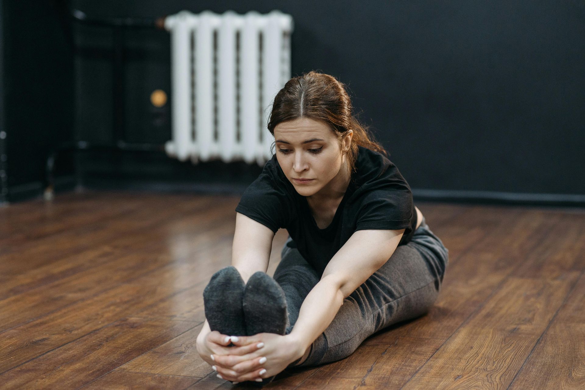 Woman in black shirt and gray pants stretches, touching her toes on a wooden floor.