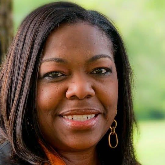 Headshot of a person with long, dark hair, wearing gold oval earrings and smiling softly against a blurred green background.