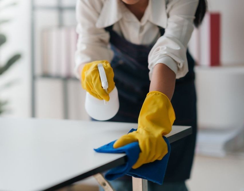 A woman wearing yellow gloves is cleaning a table with a cloth and spray bottle.
