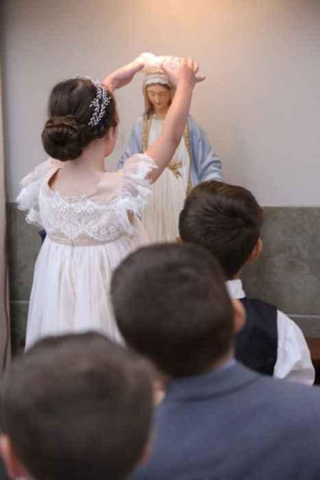 A girl in a white dress is holding a bible in front of a statue of mary