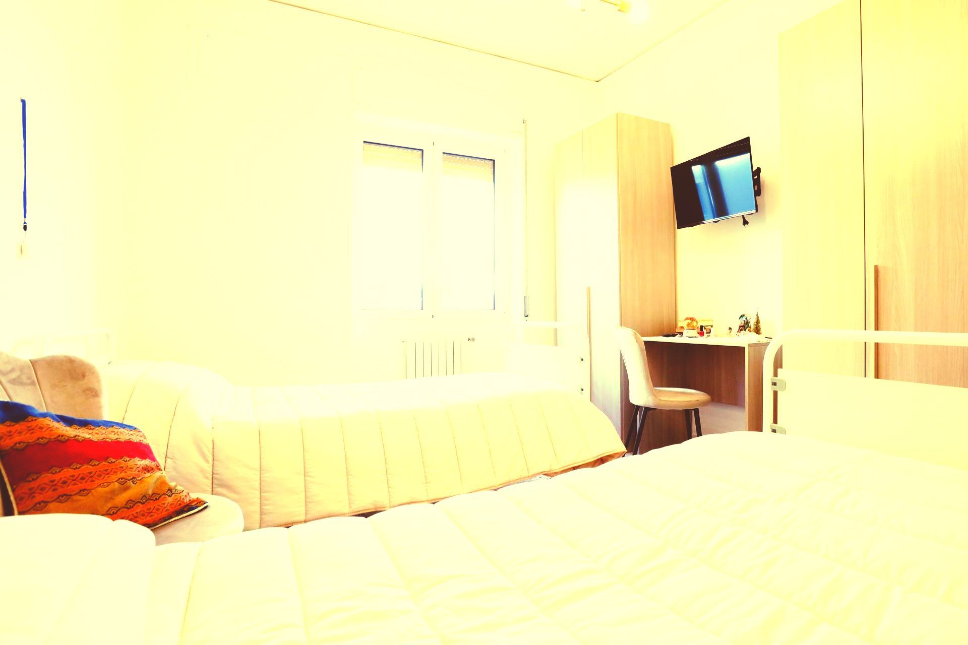 Camera da letto con due letti, scrivania, TV e armadio. Illuminazione calda e giallastra.