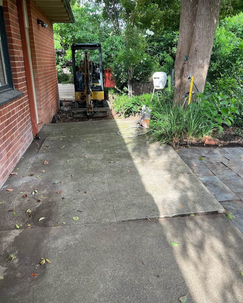 A Small Excavator is Parked in a Driveway Next to a Brick Building — CLF Landscaping In Hawker, ACT