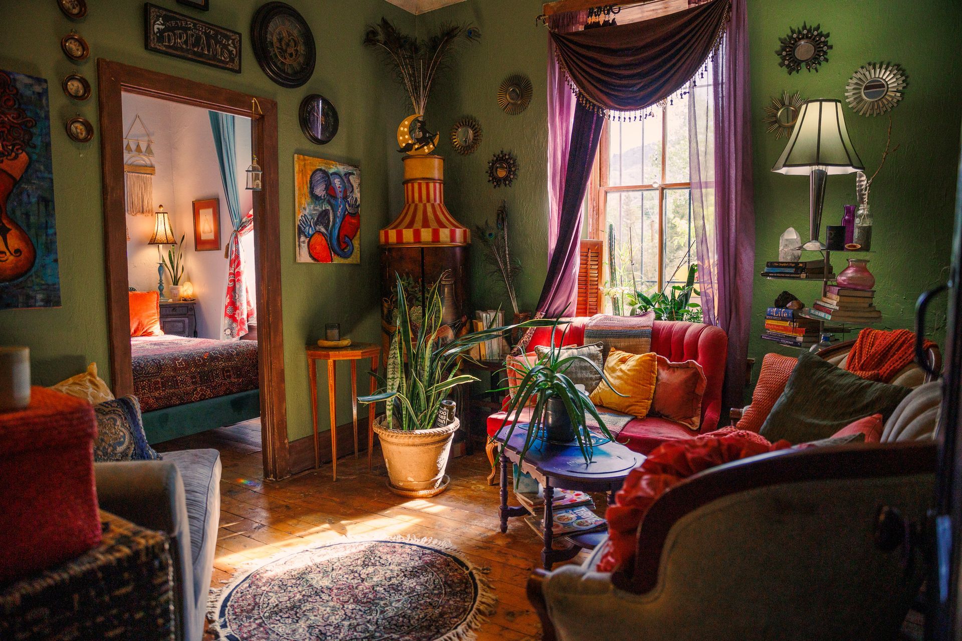 Bohemian living room with green walls, colorful furniture, multiple plants, a large window, and an open doorway to a bedroom.