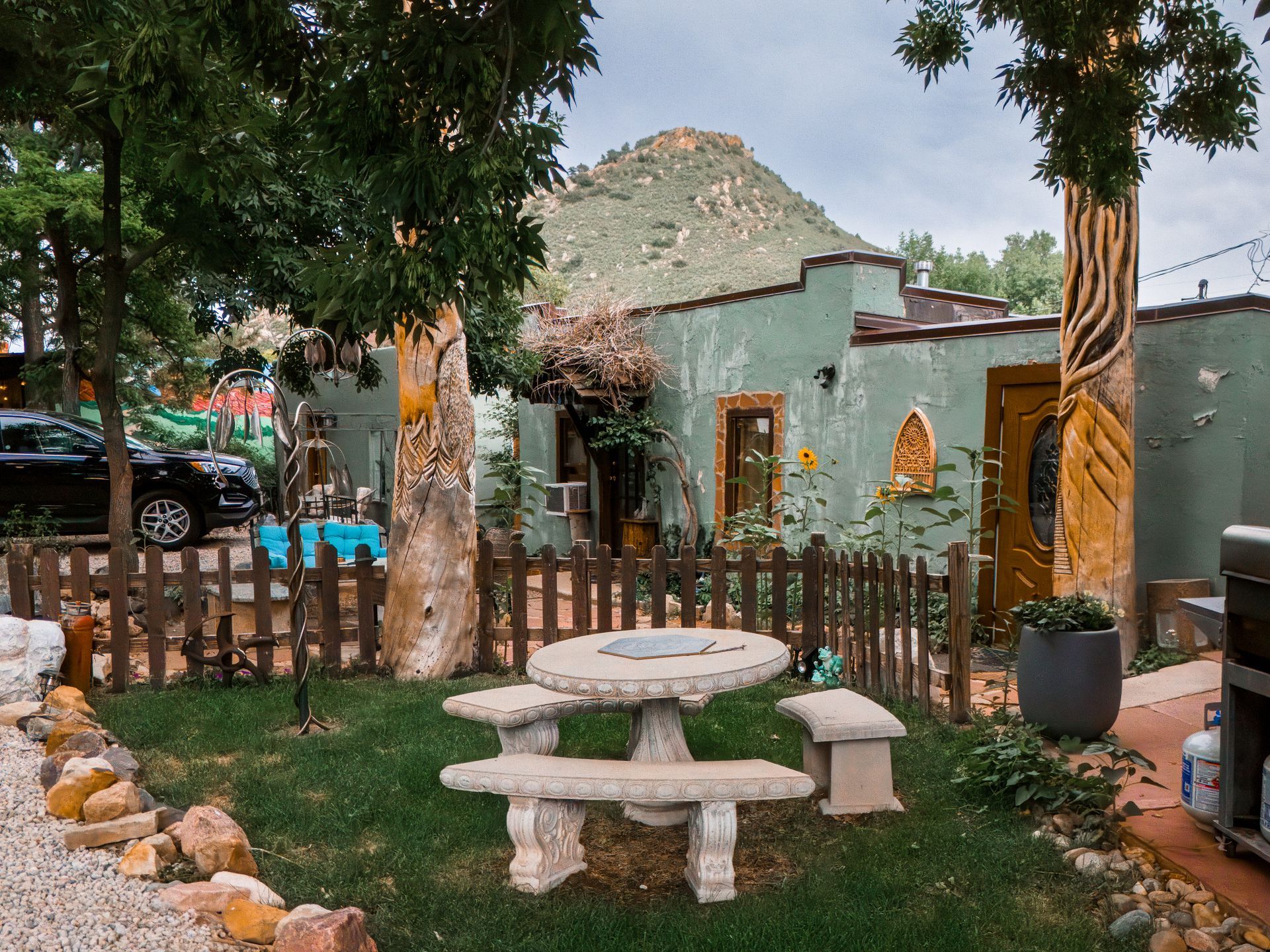 A garden area with a stone table and benches in front of a green stucco house with carved trees, near a desert mountain.