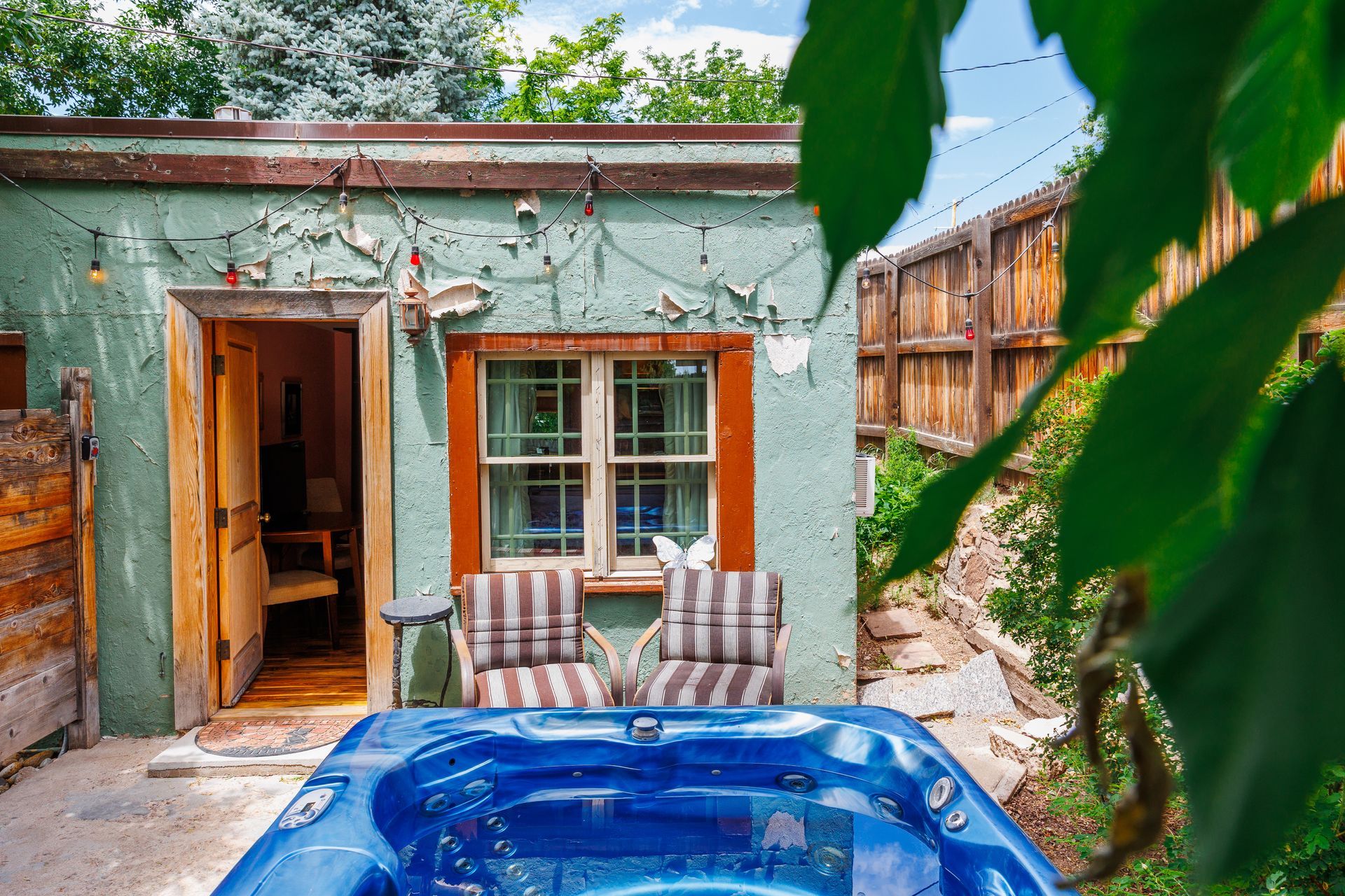 A blue hot tub sits on a stone patio in front of a weathered green building with an open door and two striped chairs.