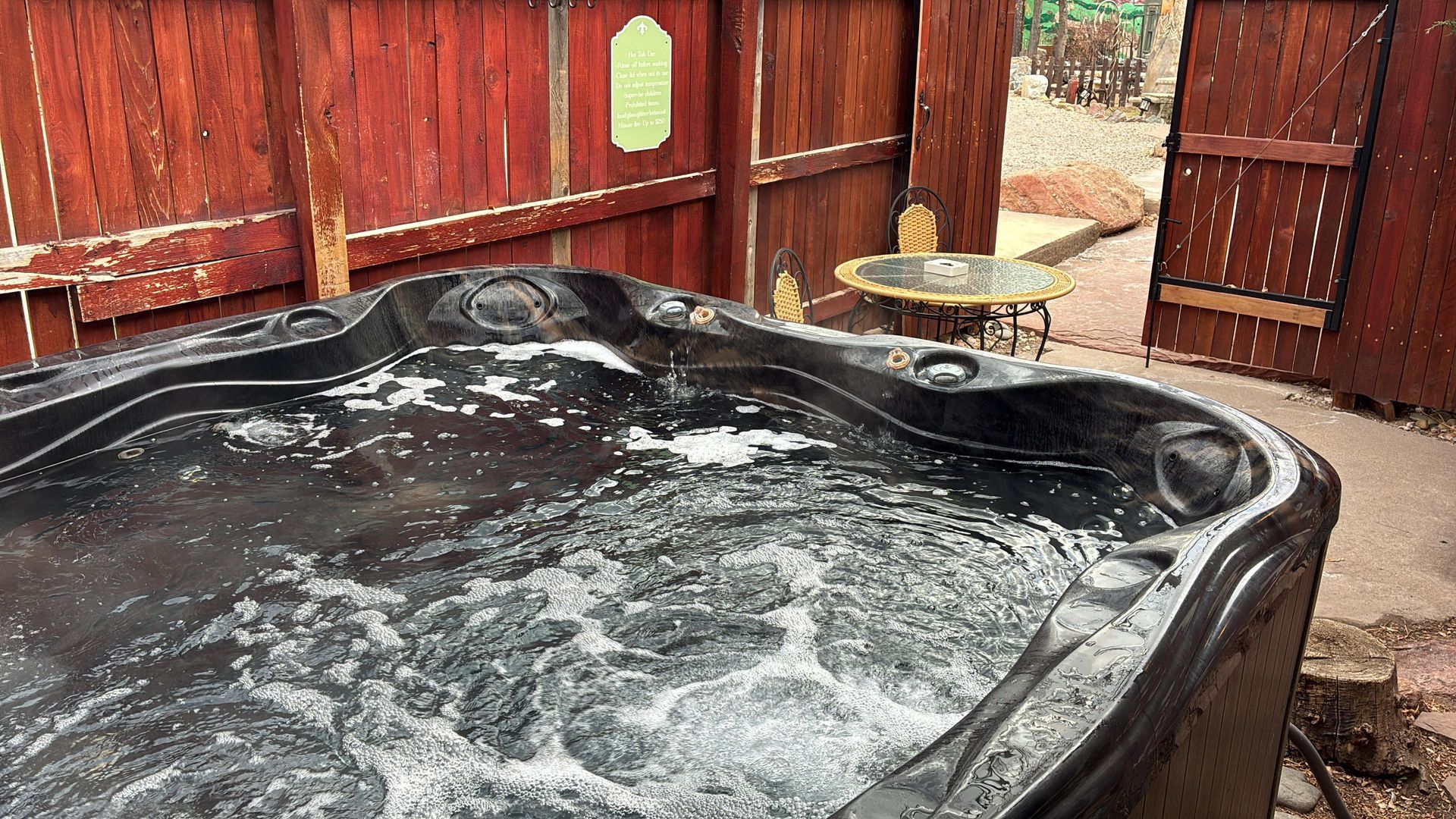 A black hot tub with bubbling water sits on a patio next to a wooden fence and a small glass-topped table.