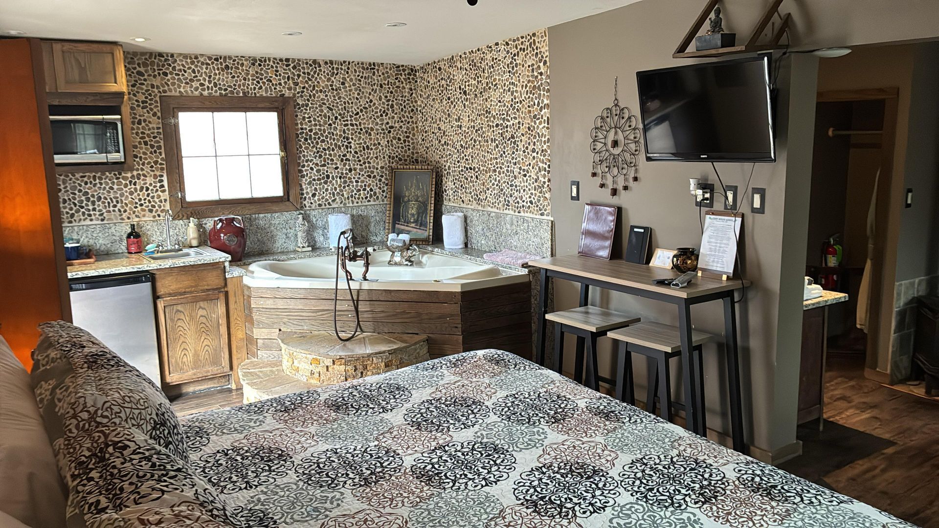 A hotel room featuring a jetted tub, a kitchenette, a bed with a patterned quilt, and a flat-screen TV on the wall.