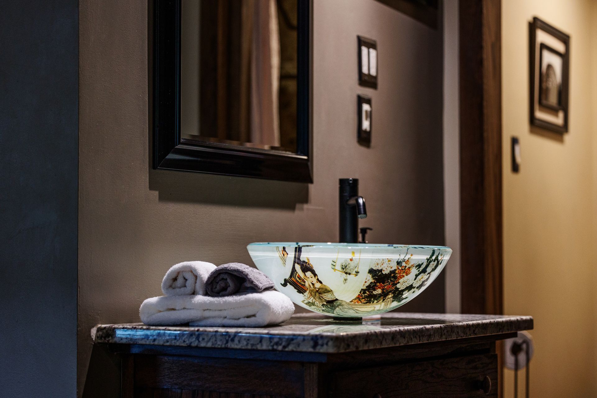 A decorative glass vessel sink sits on a stone vanity with folded towels, beneath a mirror in a dimly lit bathroom.
