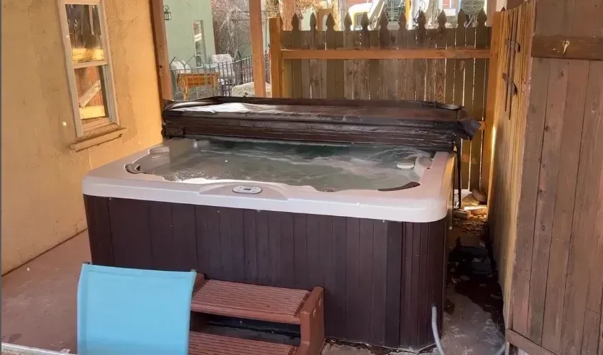 A dark-paneled rectangular hot tub with a partial cover on a porch, featuring brown steps and a light blue chair nearby.