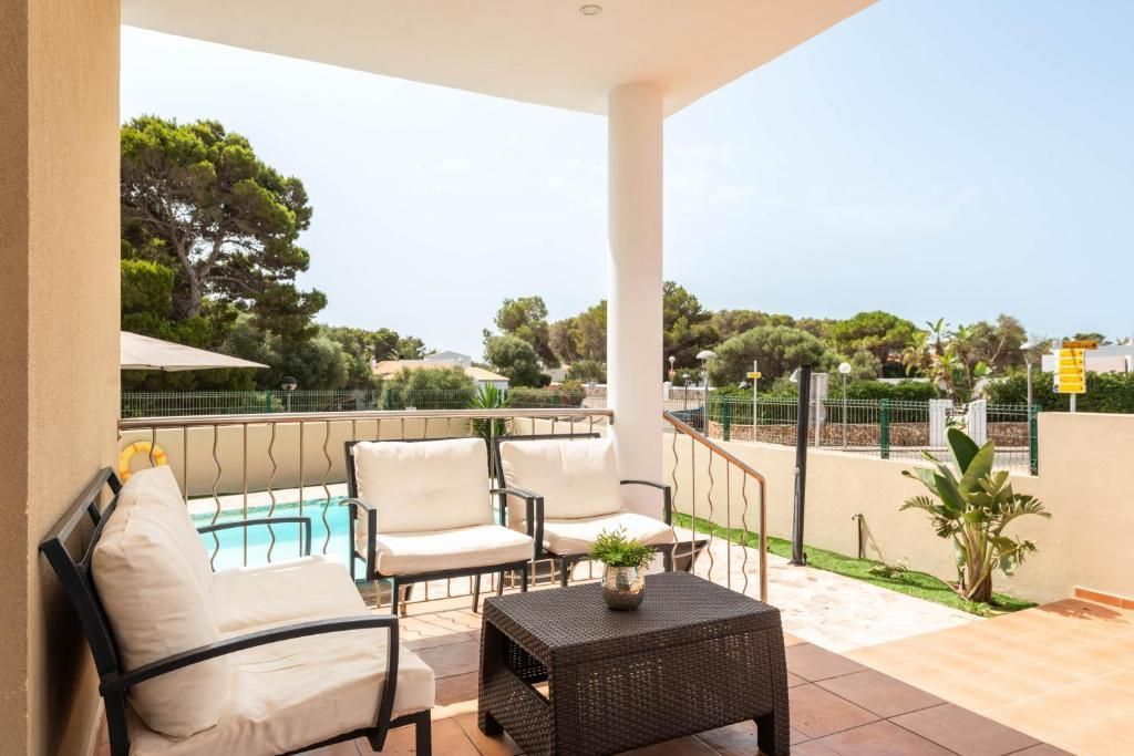 Patio con sillas blancas y una mesa marrón con vista a una piscina y vegetación.