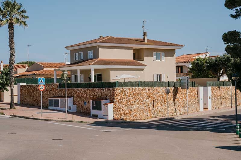 Casa de dos pisos de color bronce con pared de piedra, seto verde y palmera en una esquina de calle soleada.