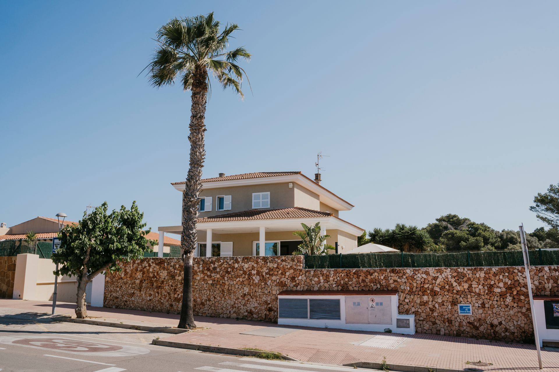 Casa con palmera frente a un muro de piedra, bajo un cielo azul.