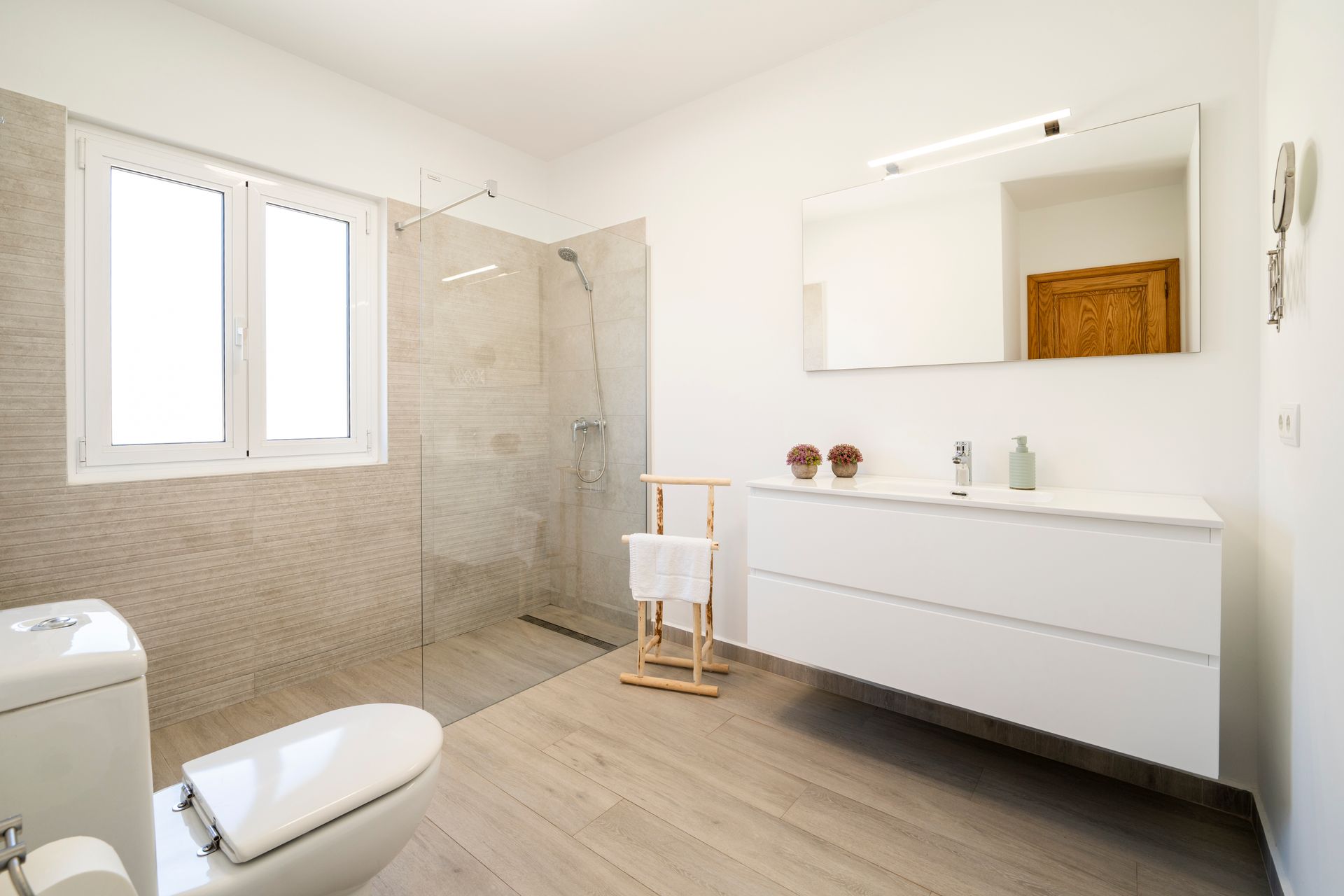 Baño moderno blanco con ducha, inodoro, lavabo y ventana. Suelo de madera clara.