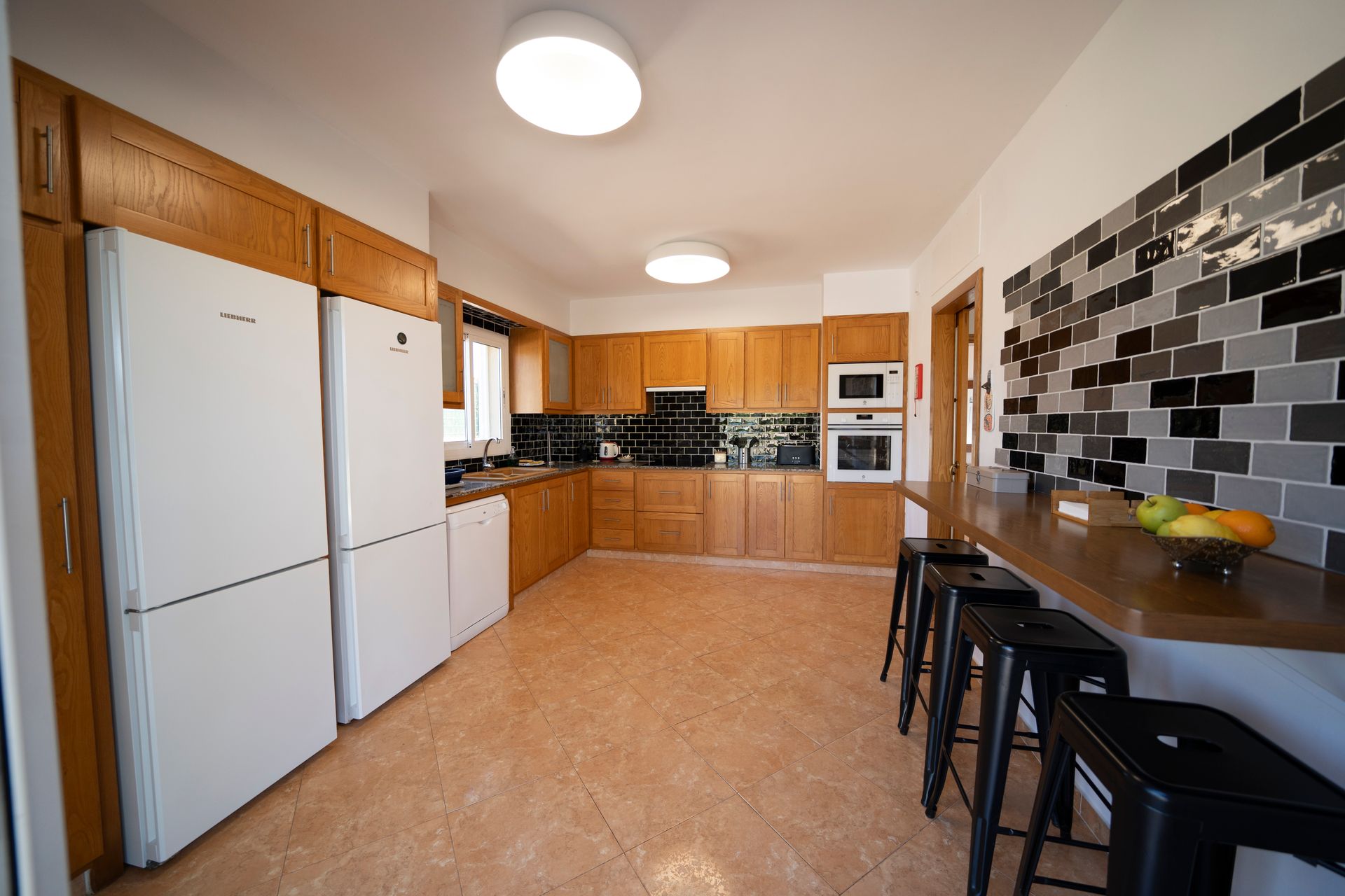 Cocina con gabinetes de madera clara, dos refrigeradores y barra de desayuno con taburetes.