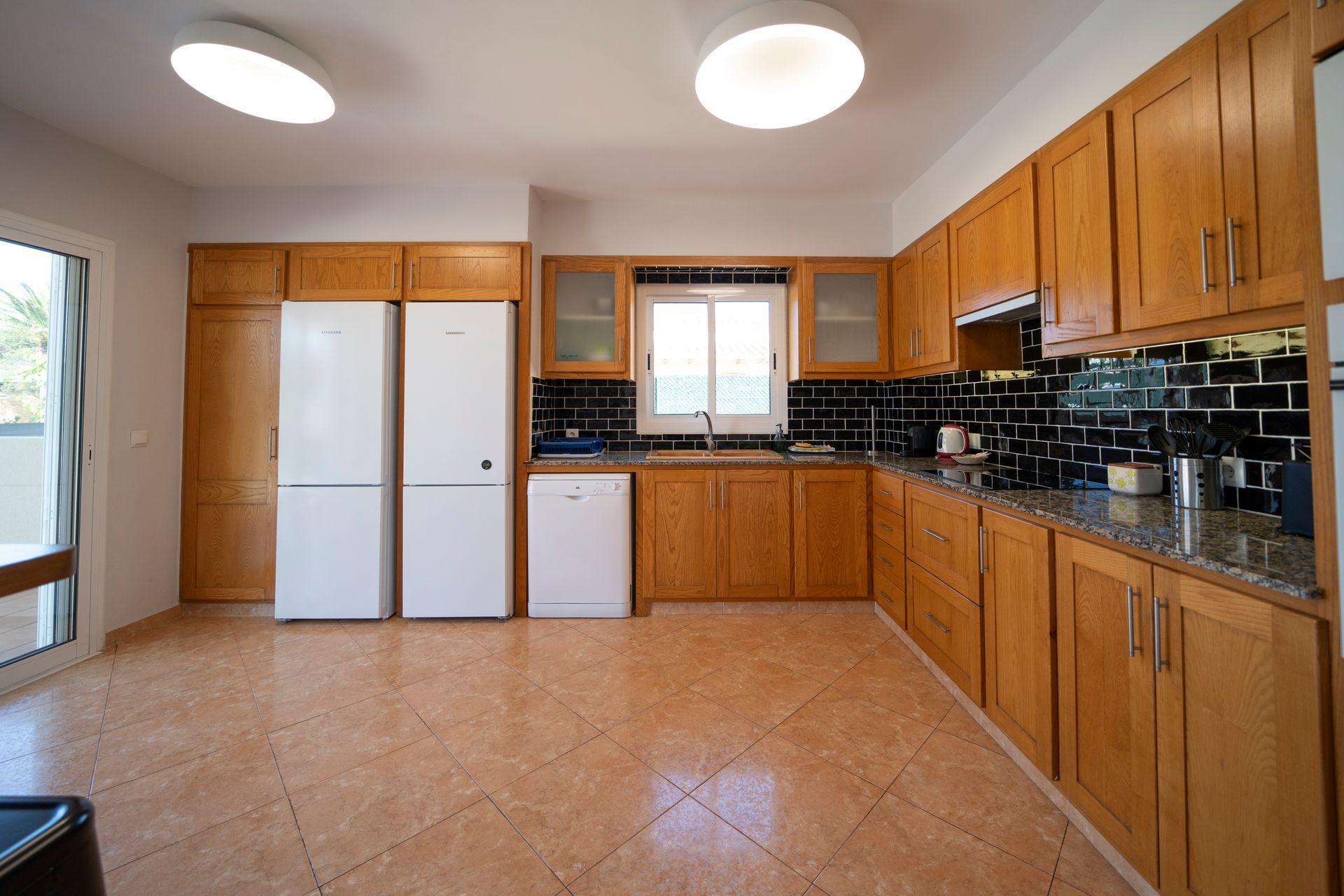 Cocina con gabinetes de madera, dos refrigeradores blancos, salpicadero de azulejos negros y piso color tostado.