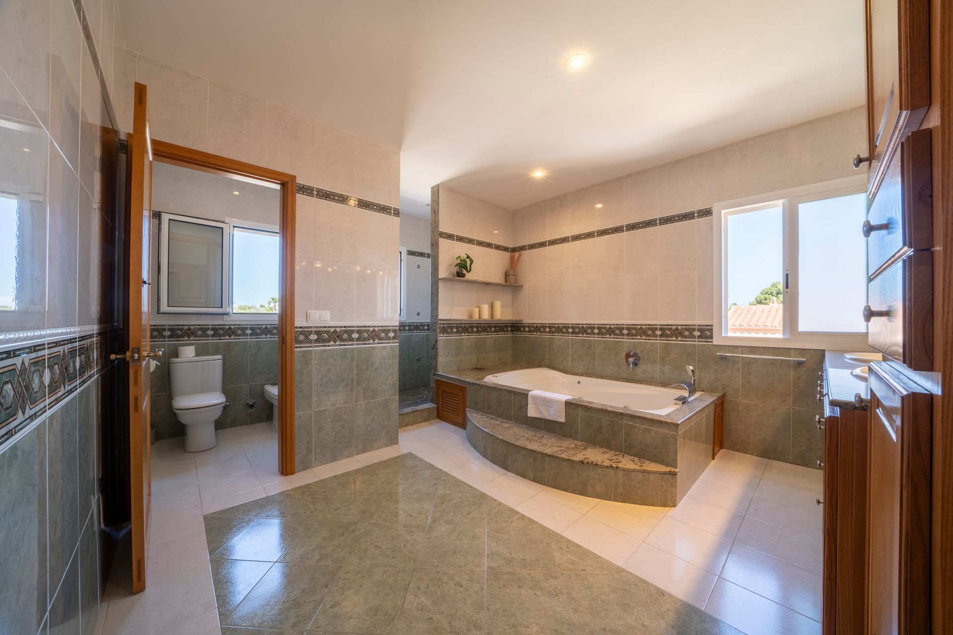 Amplio baño con jacuzzi, inodoro y amplios ventanales. Paredes claras, muebles de madera y suelo de baldosas.