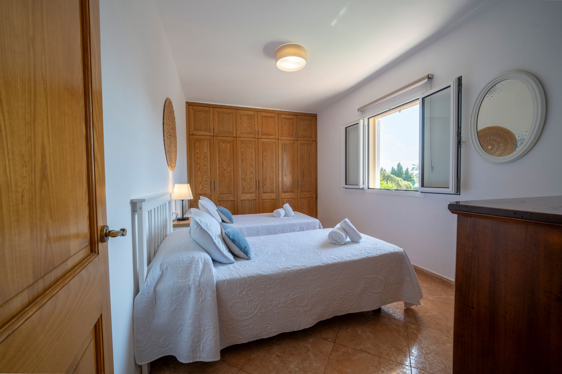 Dormitorio con dos camas individuales, ropa de cama blanca, armario y puerta de madera, ventana con mucha luz solar.