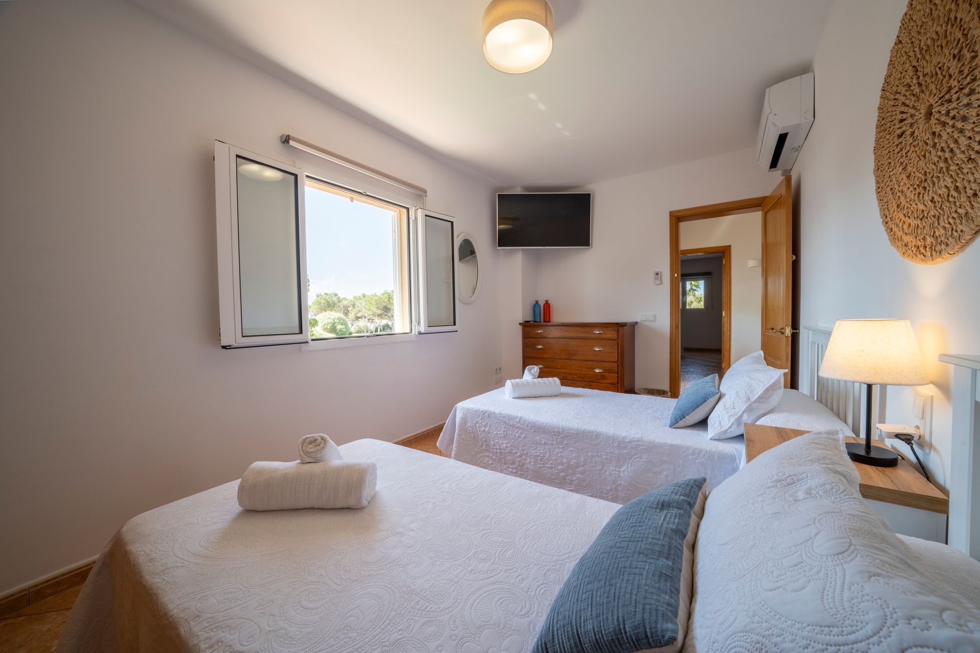 Dormitorio con dos camas individuales, cómoda, ventana, televisión pequeña y puerta abierta.