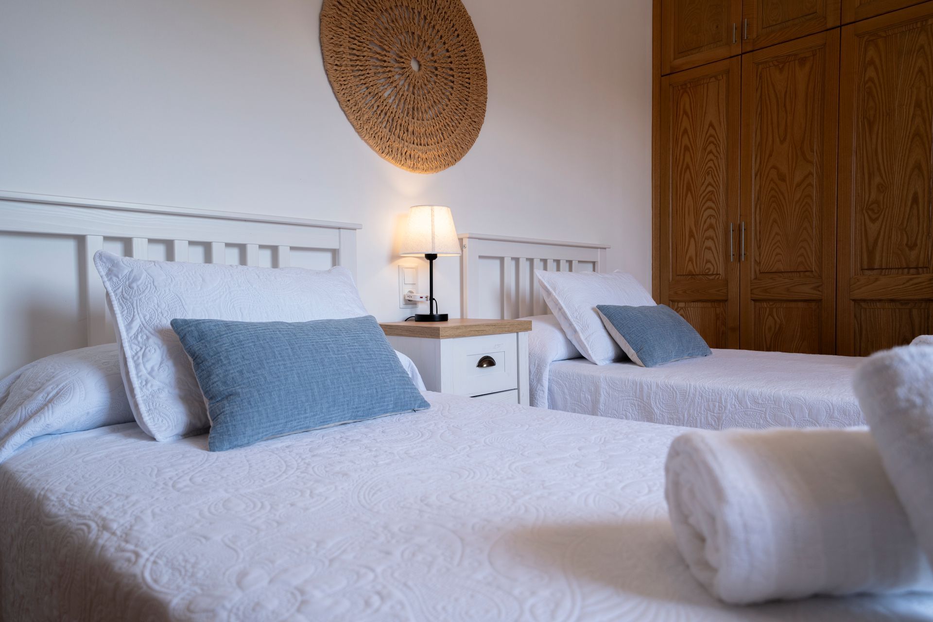 Dos camas individuales con ropa de cama blanca y almohadas azules, mesita de noche y decoración de pared tejida.
