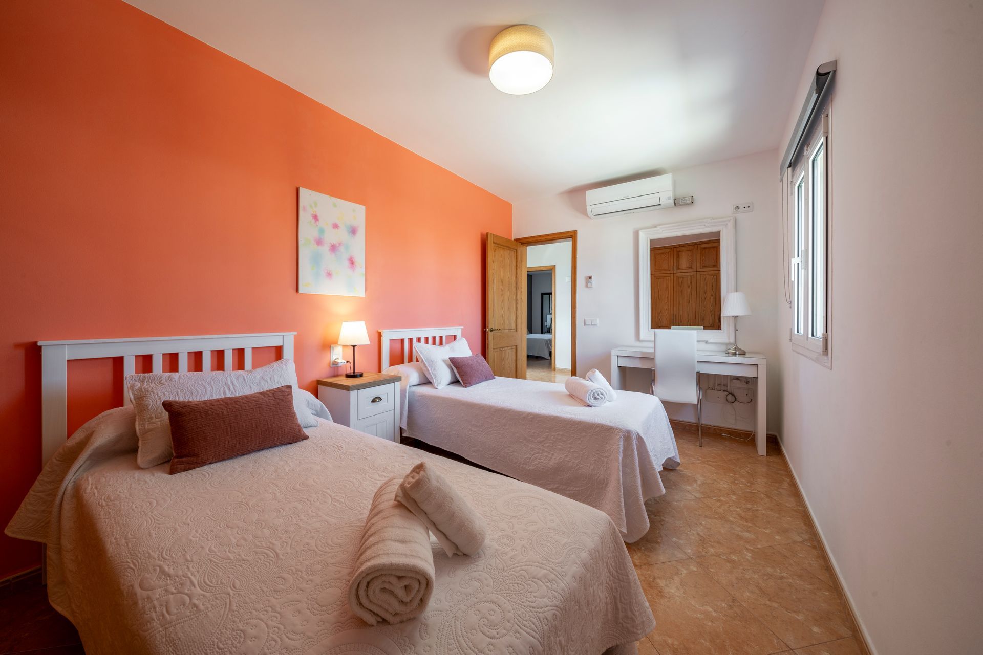 Dos camas en un dormitorio con una pared naranja. Mesillas blancas con lámparas.