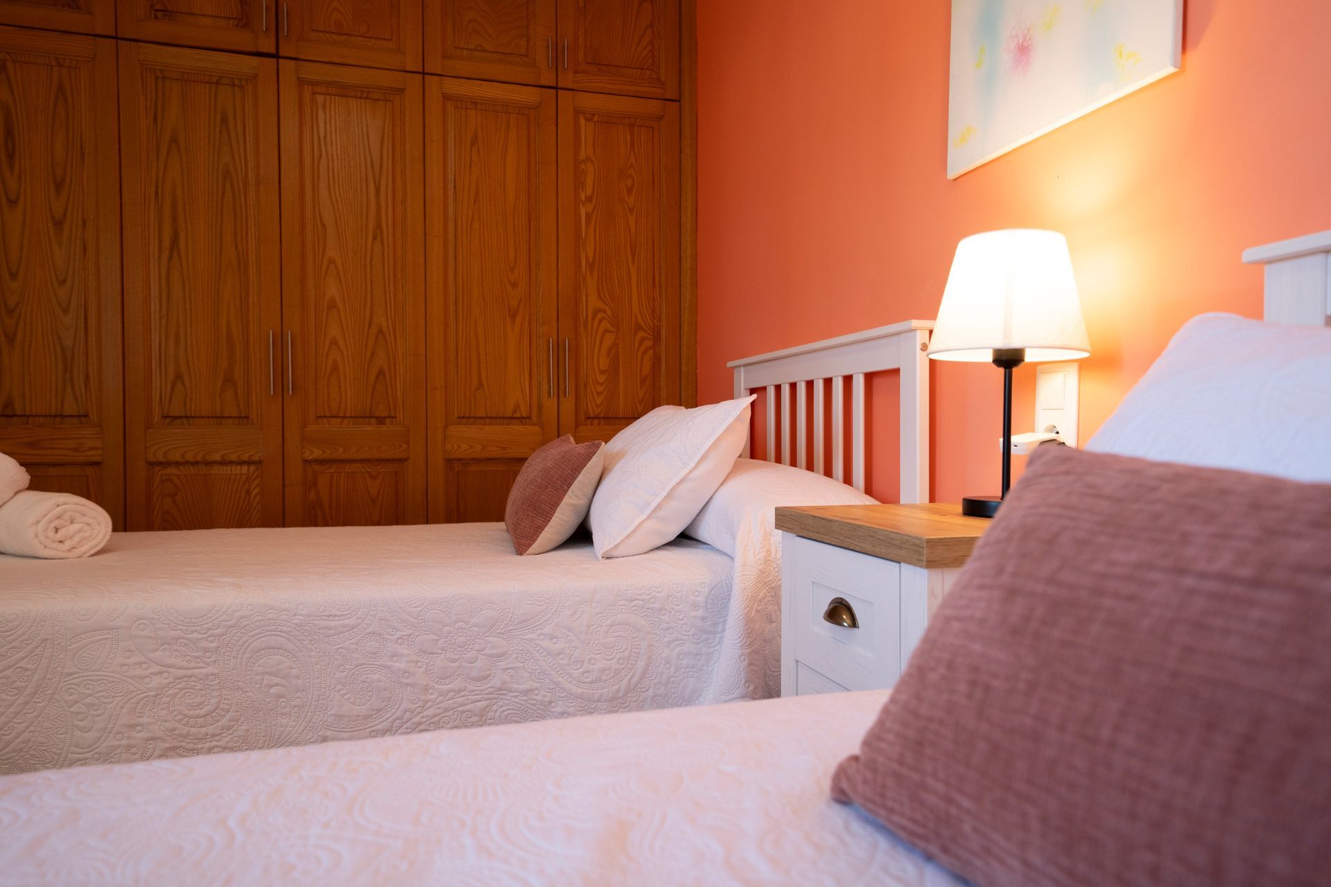 Dormitorio con dos camas individuales, armario de madera, paredes de coral, mesa de noche y lámpara.