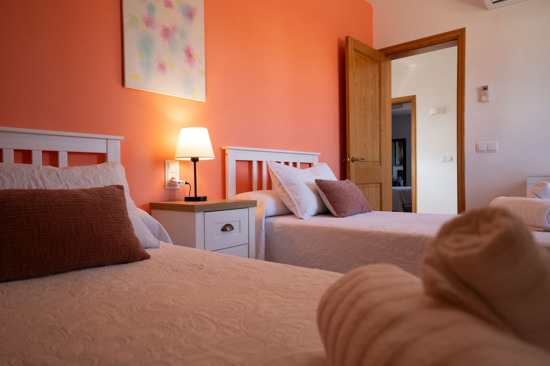 Dos camas individuales en una habitación de color melocotón, con mesita de noche, lámpara y puerta abierta.