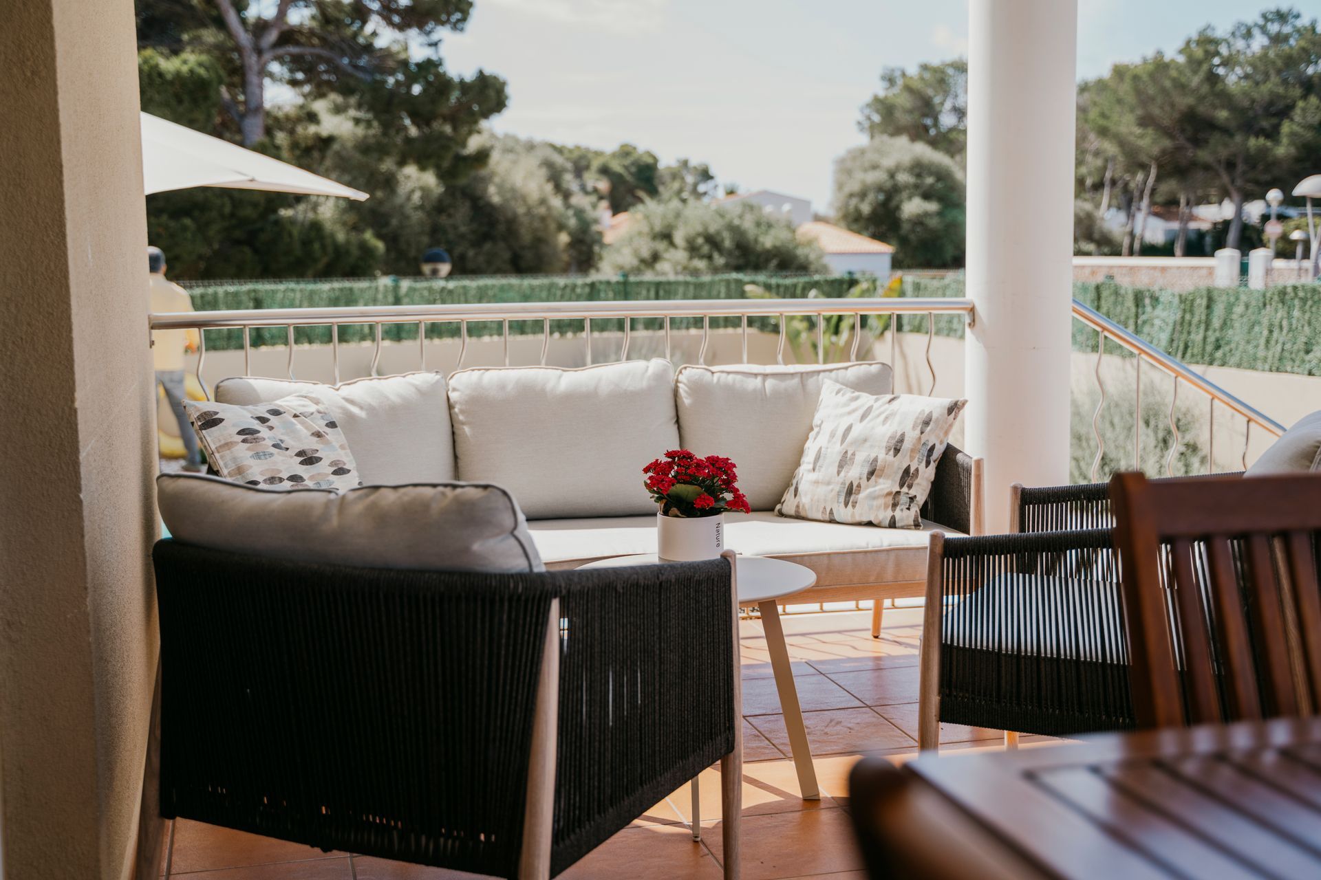Patio al aire libre con sofá acolchado, sillas, mesa pequeña y vegetación al fondo.