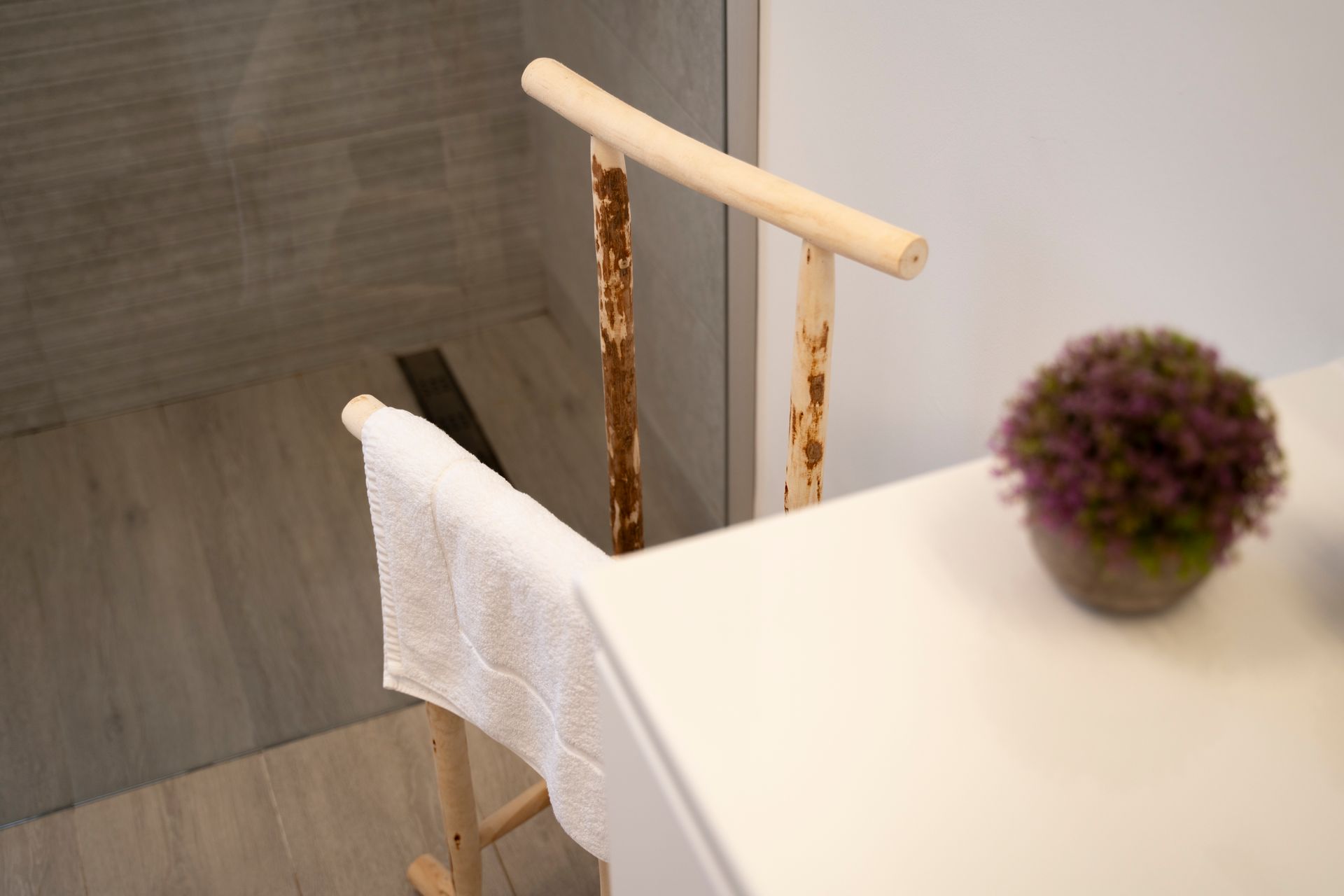 Toallero de madera con toalla blanca, junto a una maceta sobre una encimera blanca. Ducha al fondo.