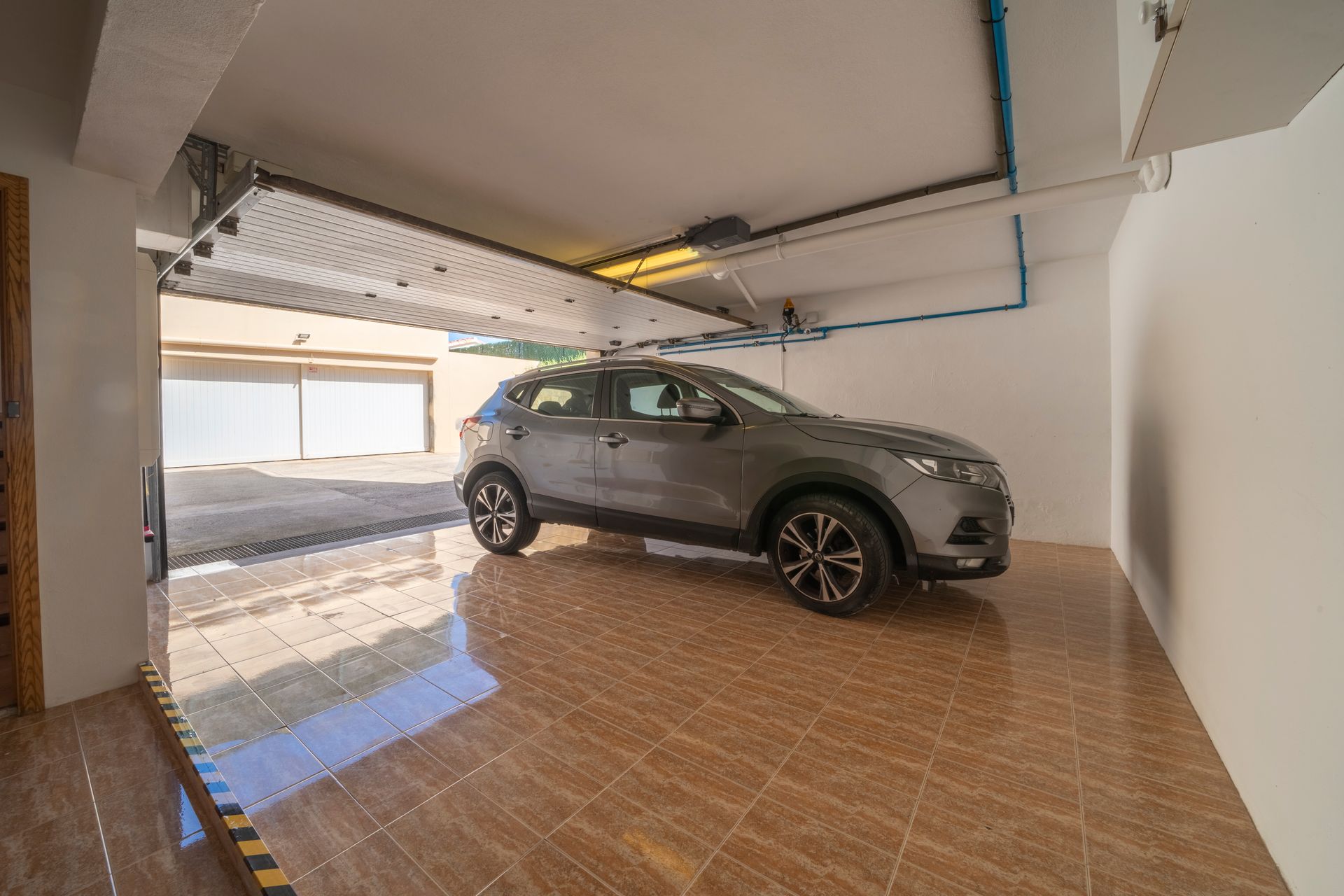 SUV gris en un garaje, suelo de baldosas, puerta abierta a la entrada. Mecanismo de la puerta del garaje visible.