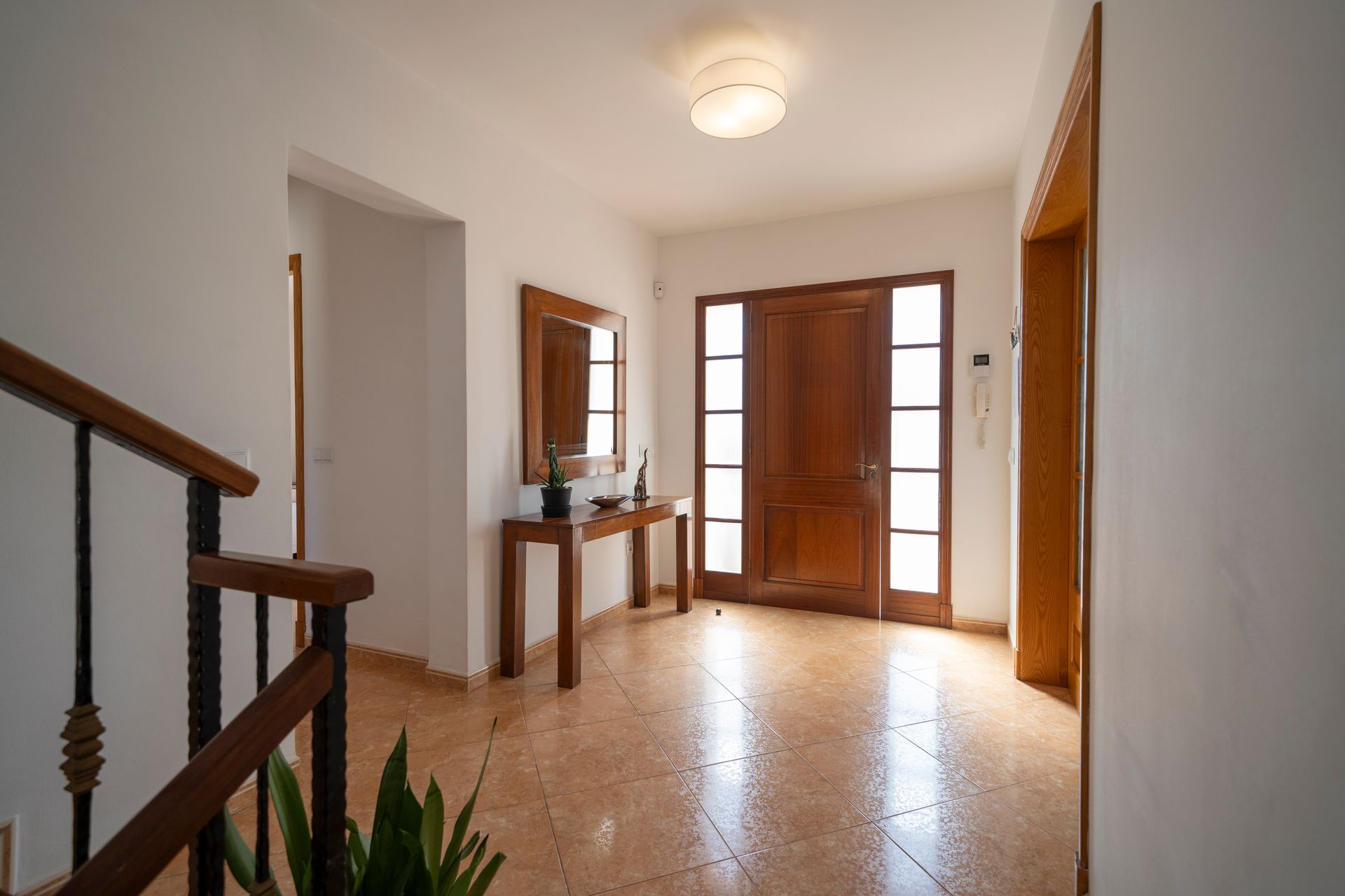 Recibidor con puerta de madera, mesa con espejo y escalera. Luminoso con suelo de baldosas.