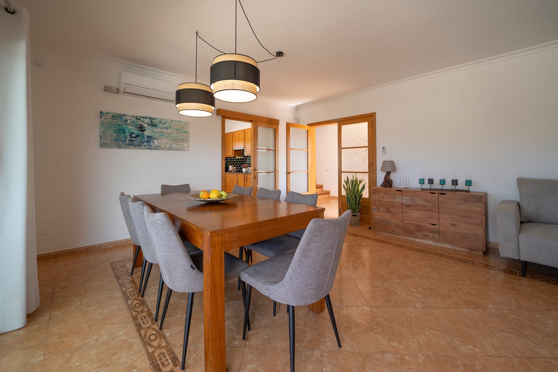 Comedor con mesa de madera y sillas grises. Iluminación decorativa y aparador.