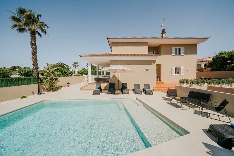 Casa con piscina, patio con camastros, sombrilla y palmeras bajo un cielo azul claro.