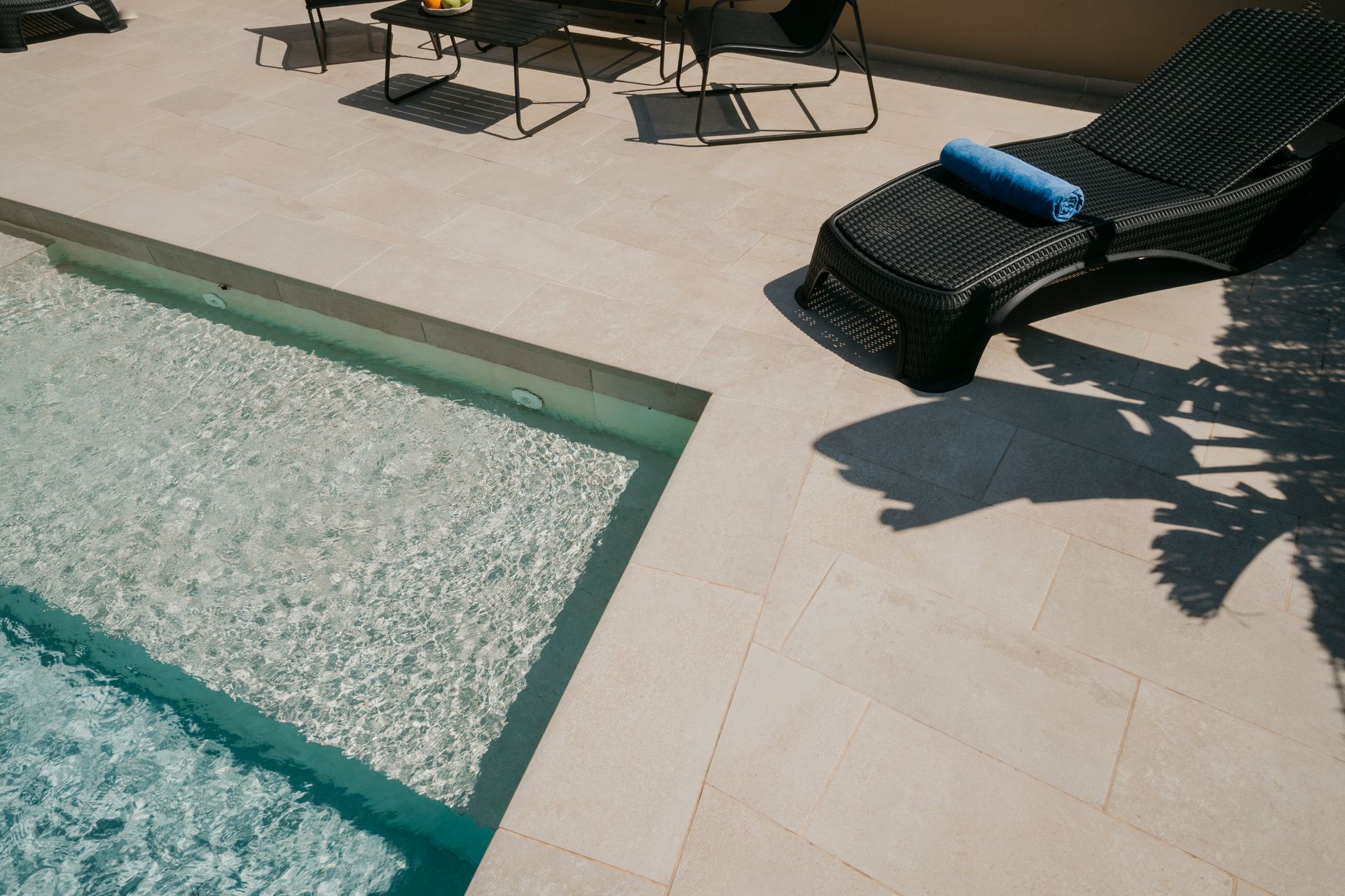 Escena junto a la piscina: piscina con agua cristalina, tumbona y muebles de patio sobre azulejos beige. Día soleado.