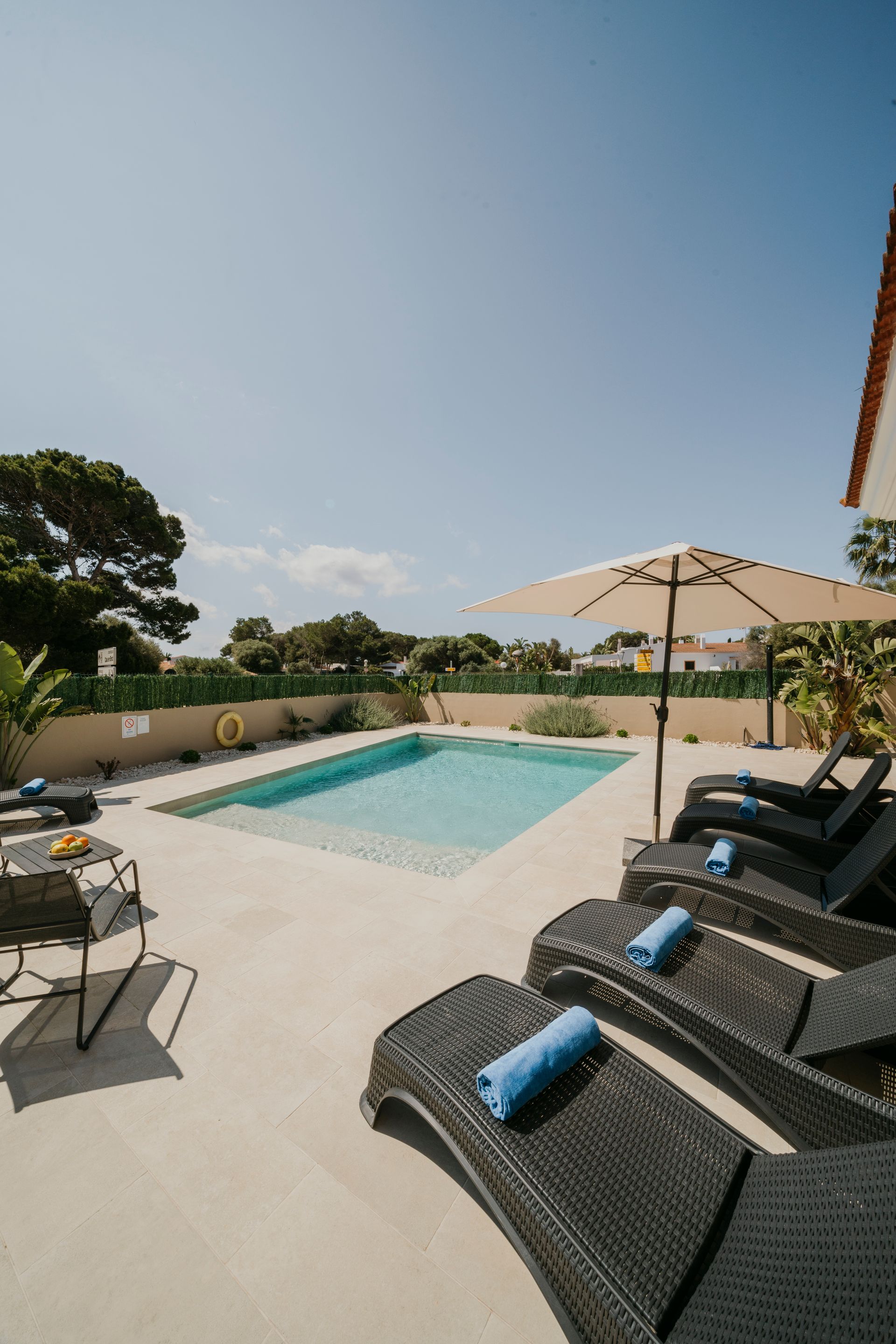 Escena junto a la piscina con tumbonas, sombrilla y un cielo azul claro.