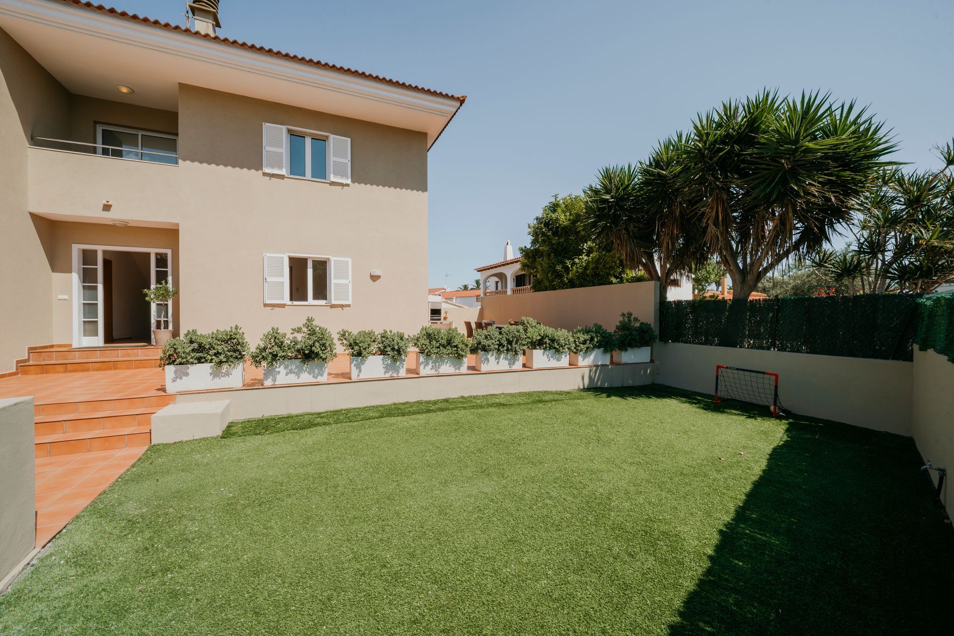 Casa de dos plantas color canela con un pequeño jardín verde, jardineras blancas y un seto. Cielo azul.