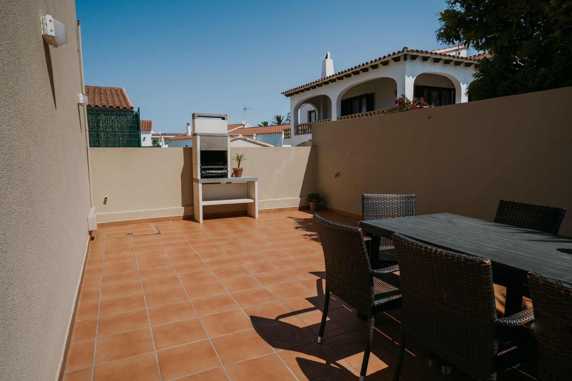 Terraza con barbacoa, mesa de comedor y sillas. Día soleado con paredes beige, azulejos marrones y una casa blanca al fondo.