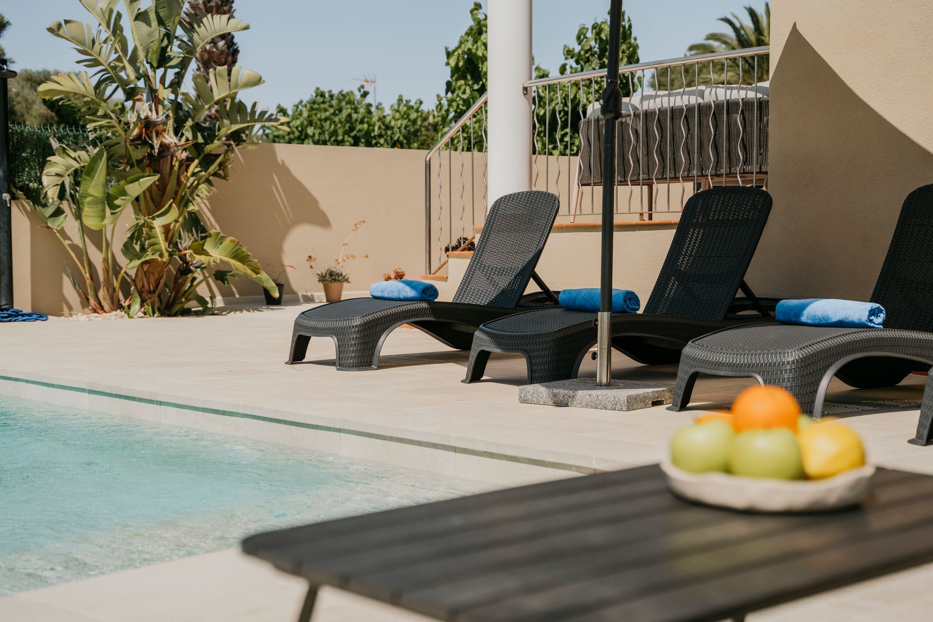 Tumbonas junto a una piscina con fruta en una mesa, día soleado.