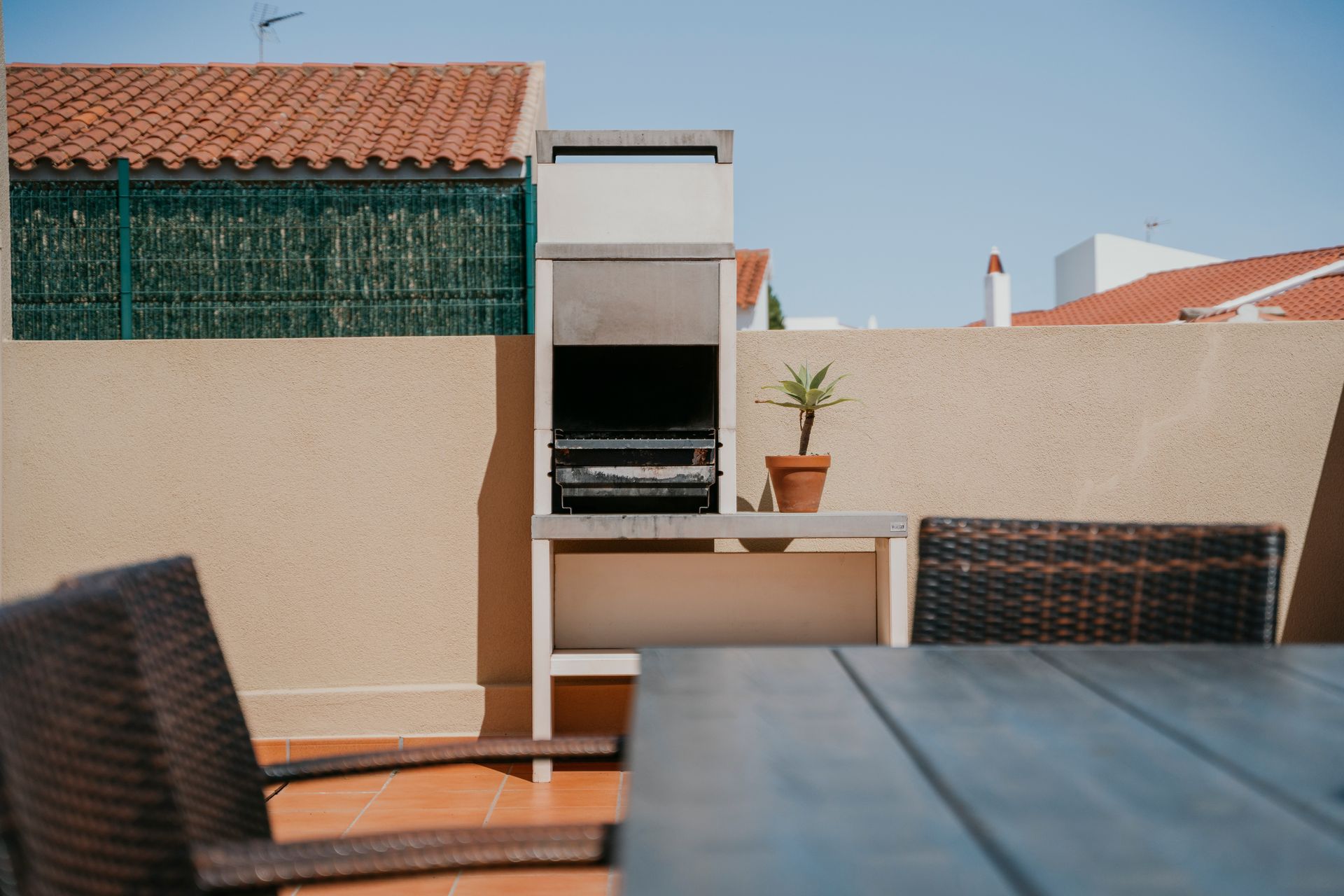 Parrilla de barbacoa en un patio soleado con mesa, sillas y una pequeña planta en maceta.