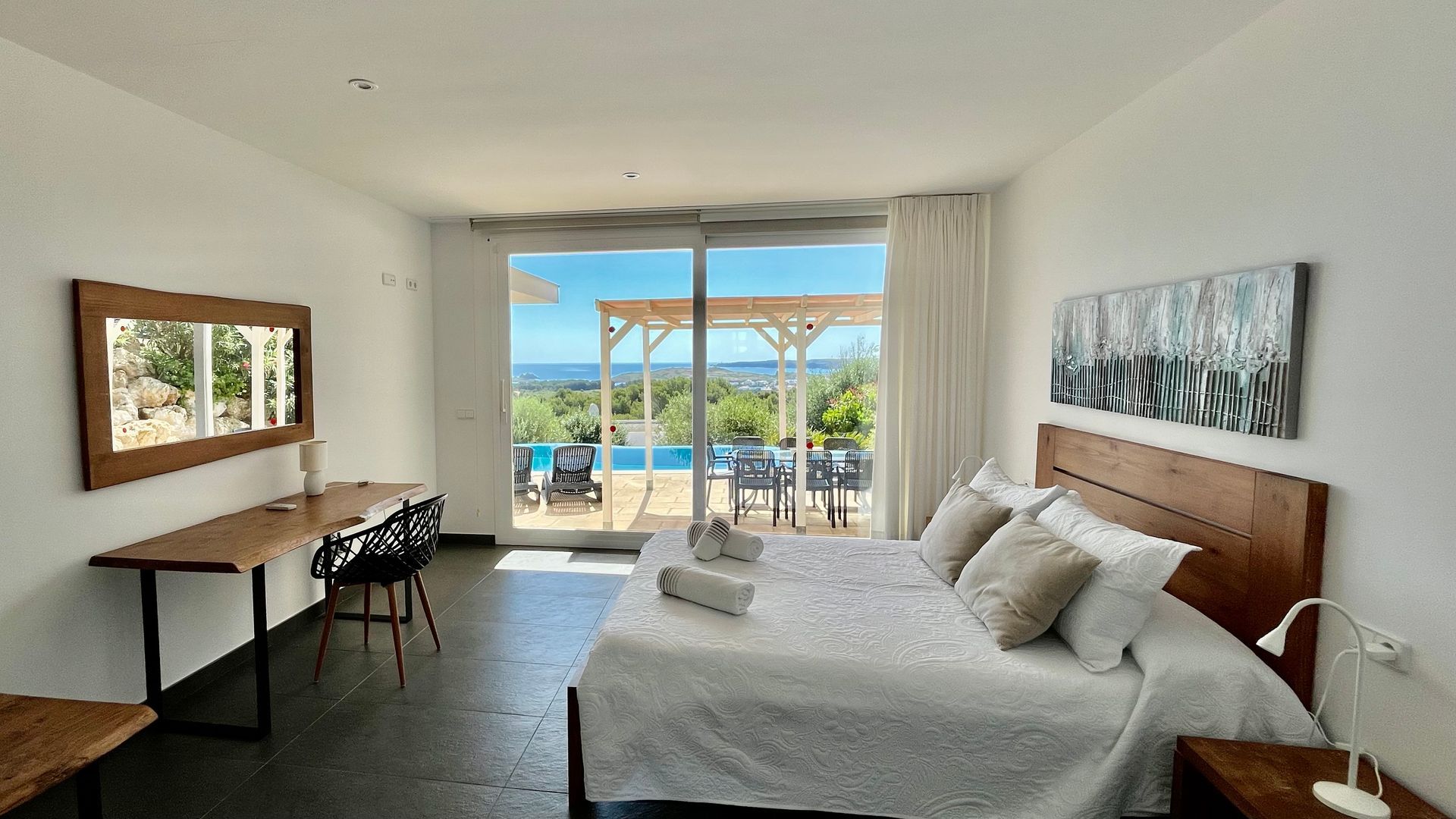 Dormitorio con gran ventanal con vistas a la piscina y al mar; paredes blancas, muebles de madera oscura.