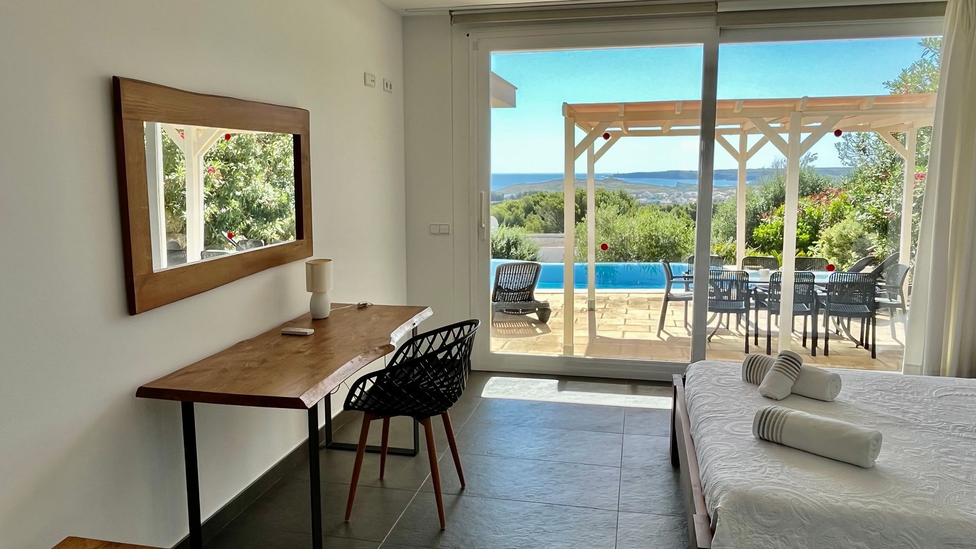 Dormitorio con escritorio, espejo y puertas abiertas a piscina exterior, vista al mar.